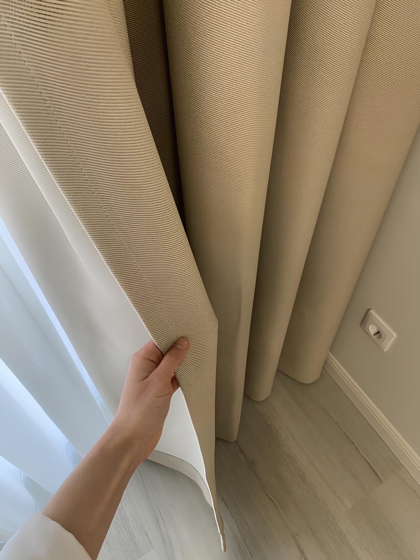 Hand holding back beige striped curtains next to sheer white curtains in a room corner.
