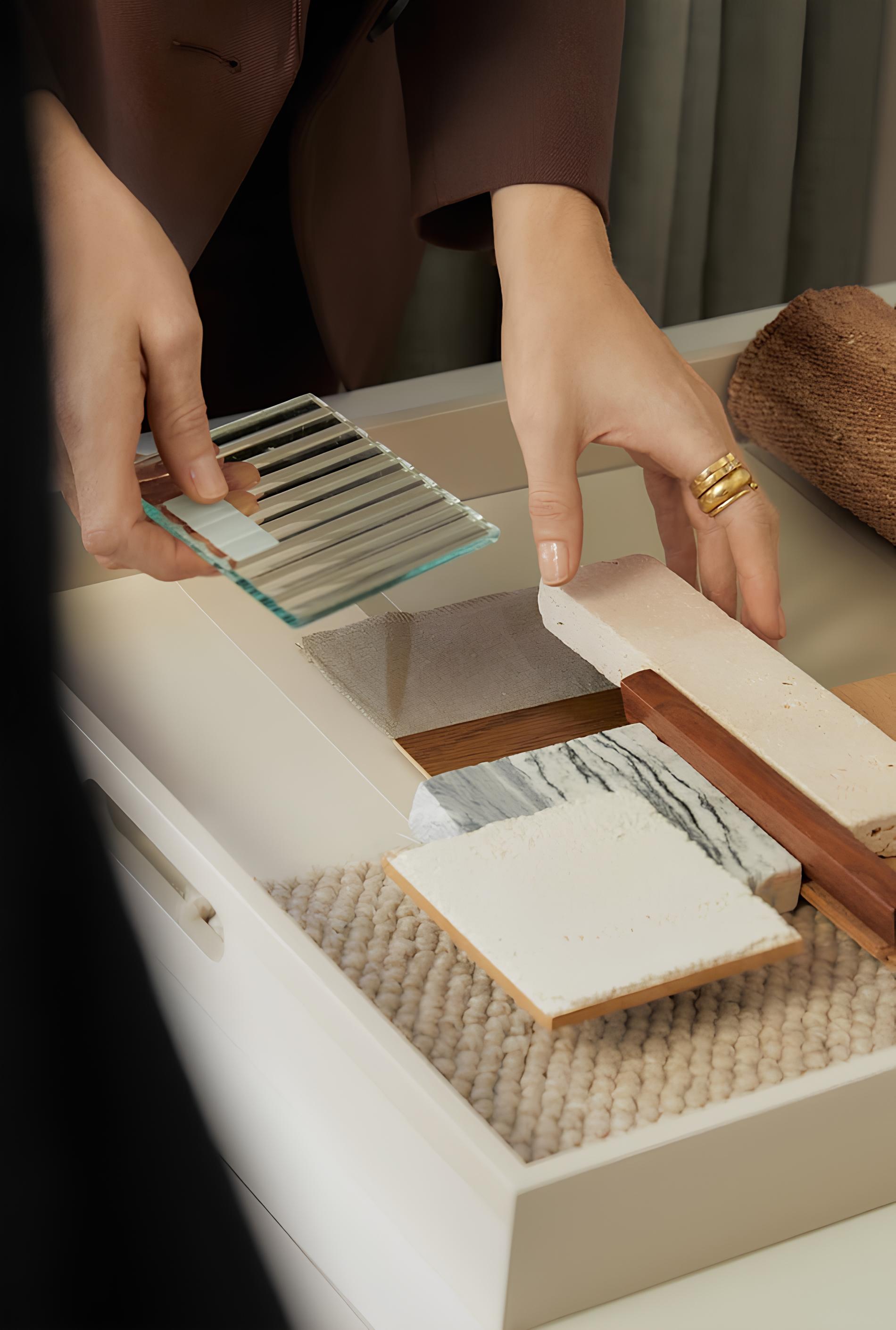 Hands arranging textured and patterned material samples including fabric, stone, wood, and glass on a surface.