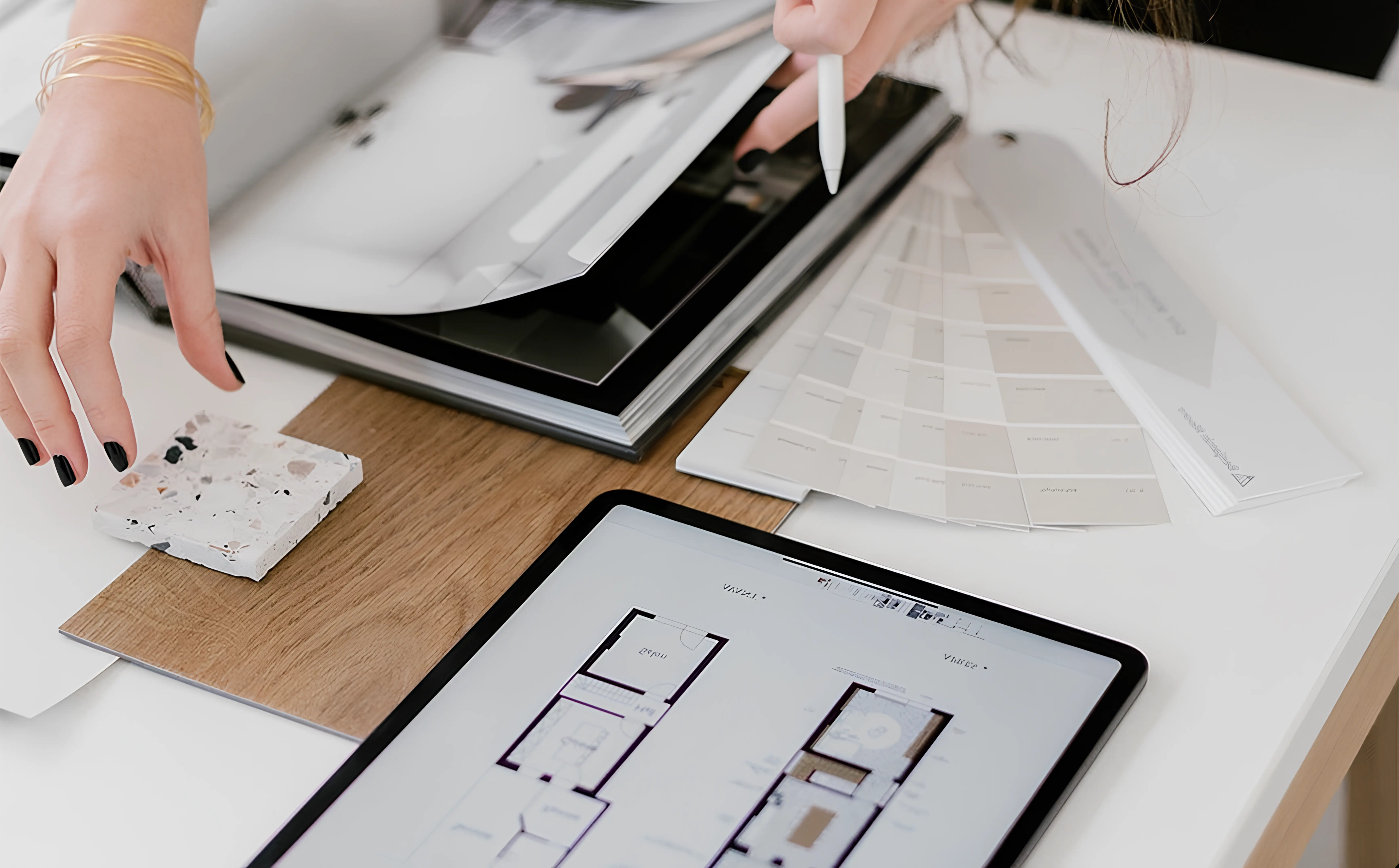 Hands with black nail polish selecting materials and floor plans on a table with tablet and color swatches.