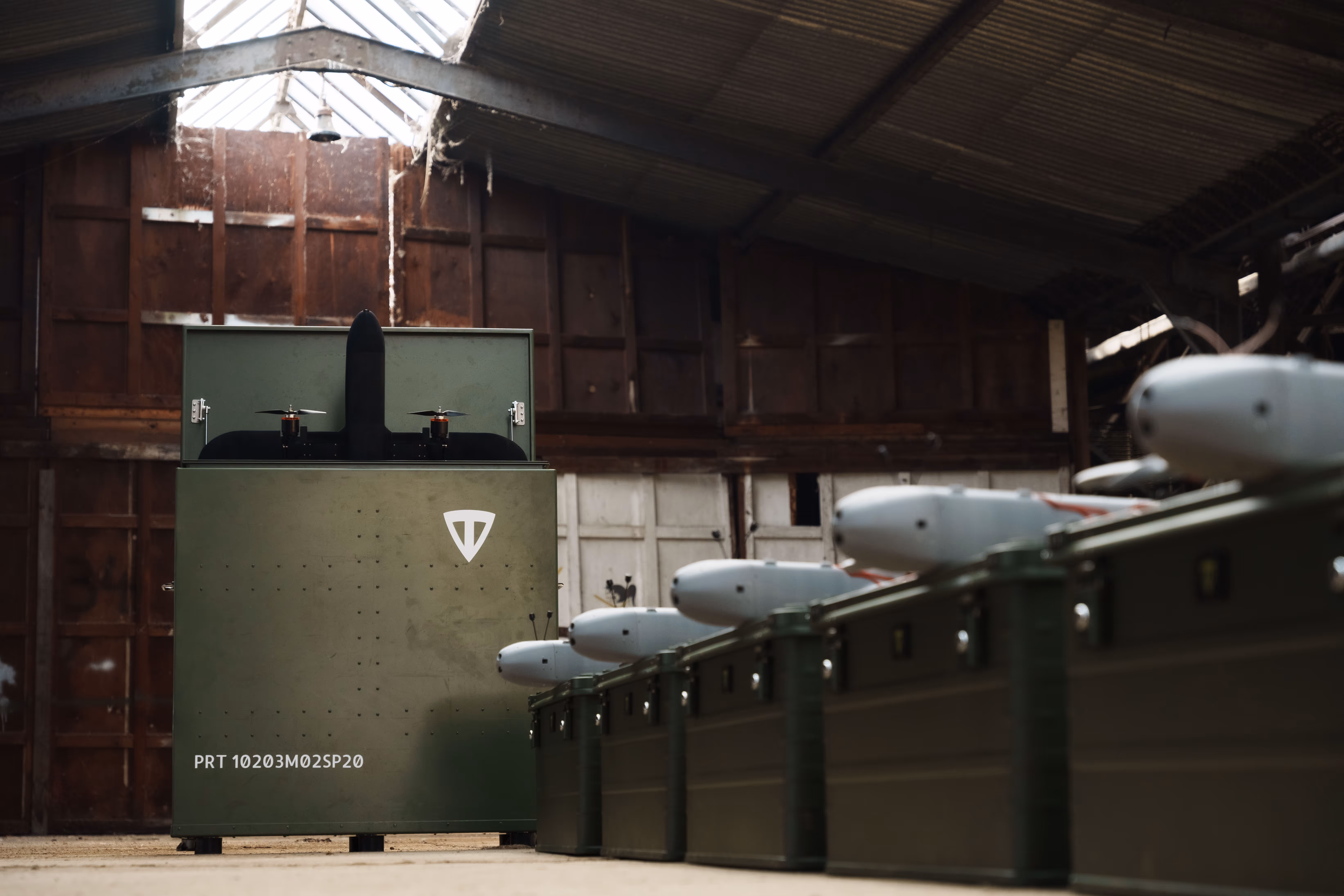 Row of olive green military-style containers with white drone parts, inside a warehouse with a large green box at the end featuring a white emblem and two small drone propellers on top.