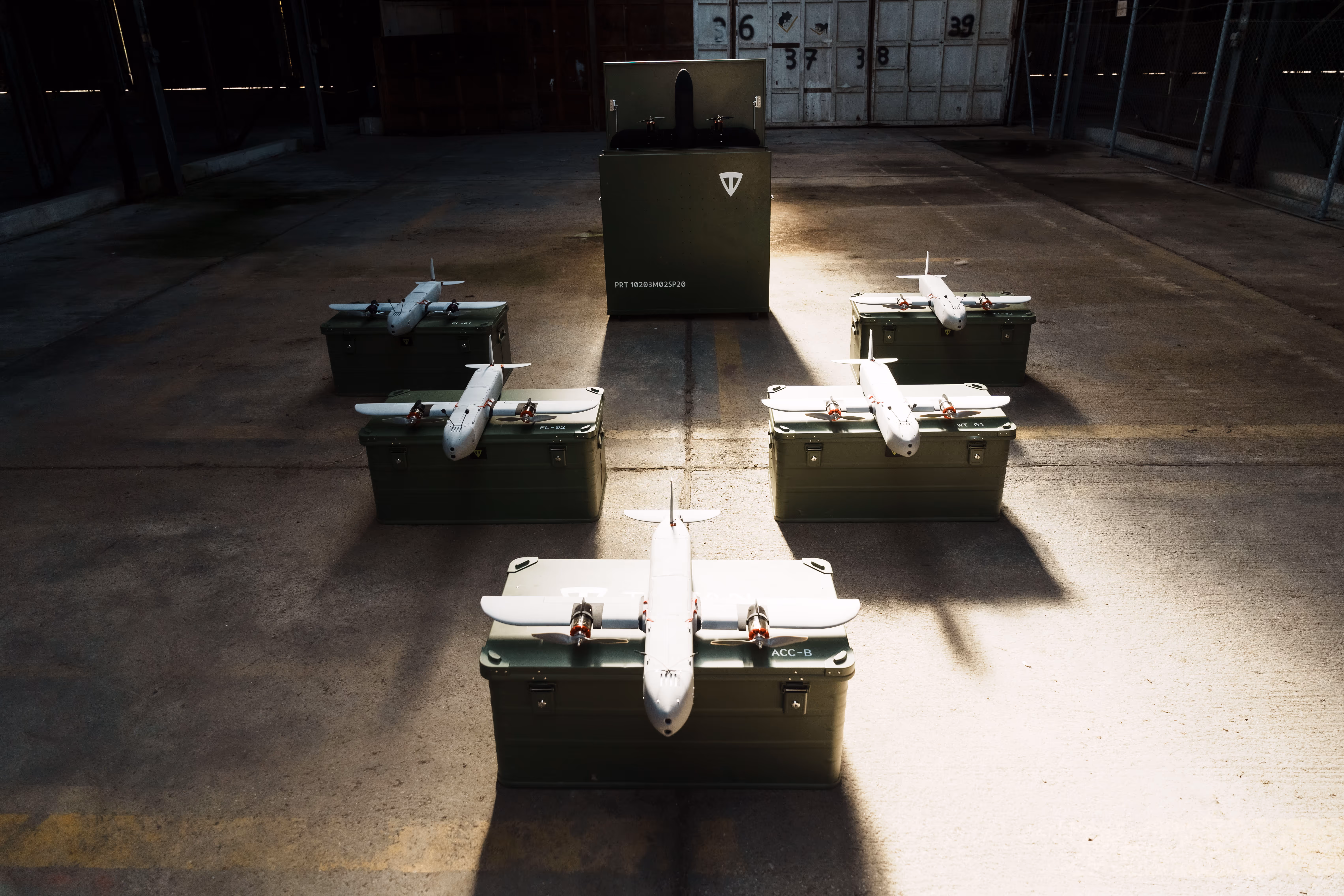 Five small white drones with dual propellers placed on green military-style storage boxes in a dimly lit industrial warehouse.