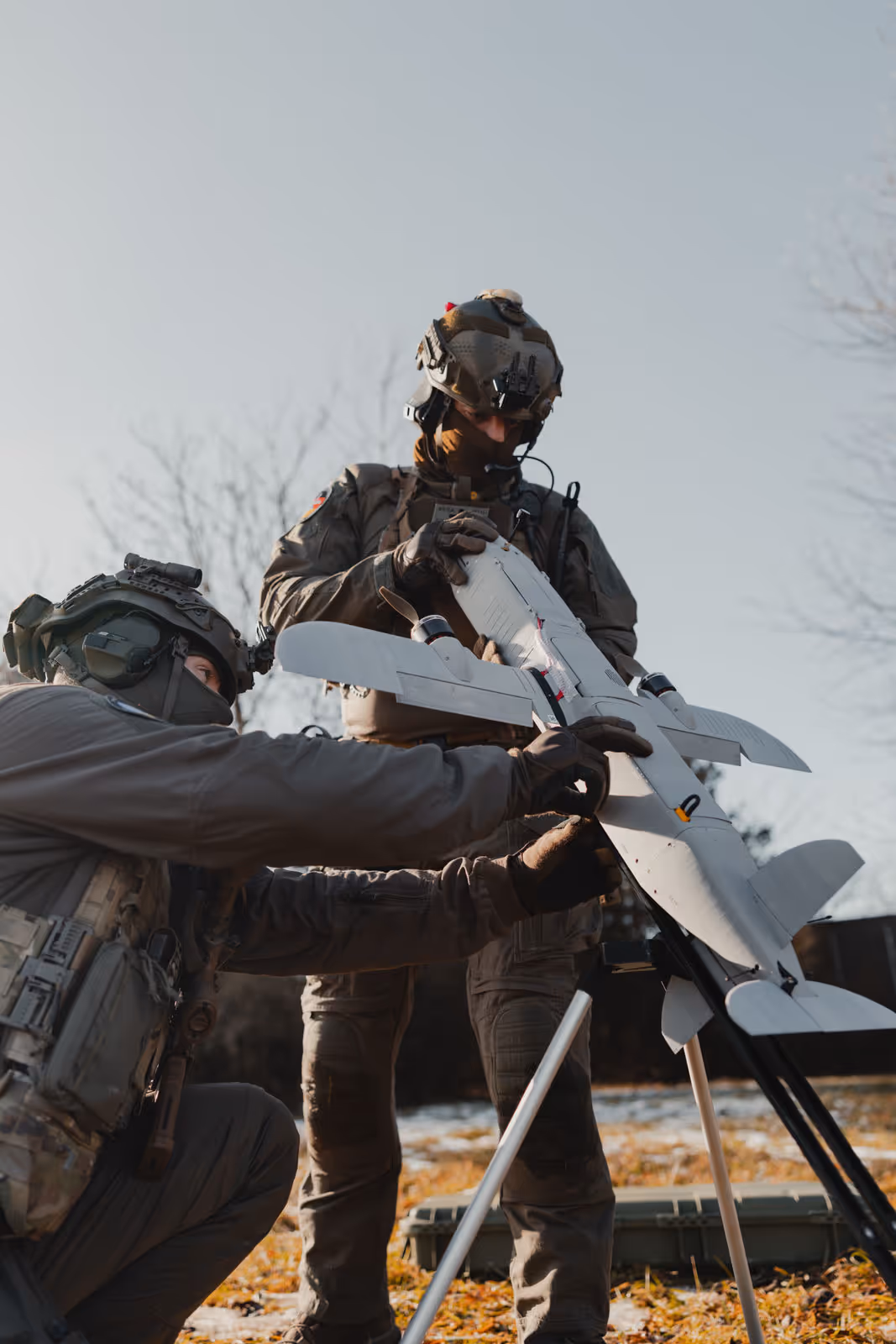 Two soldiers in tactical gear preparing a small drone launcher outdoors on a grassy area.