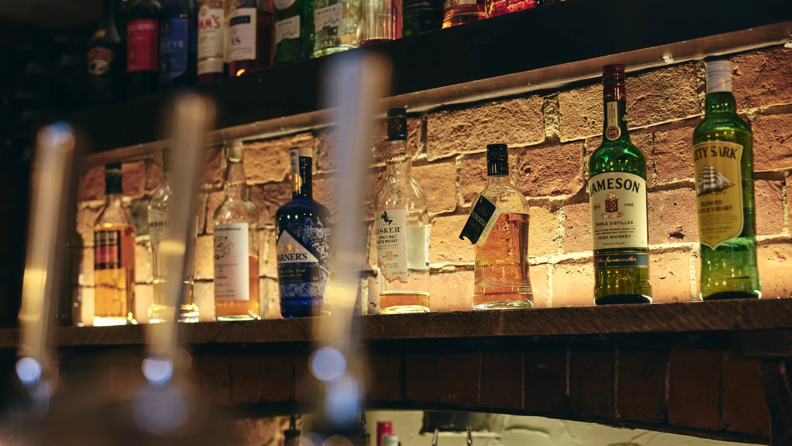 Shelf of various whiskey bottles illuminated against a brick wall in a bar.