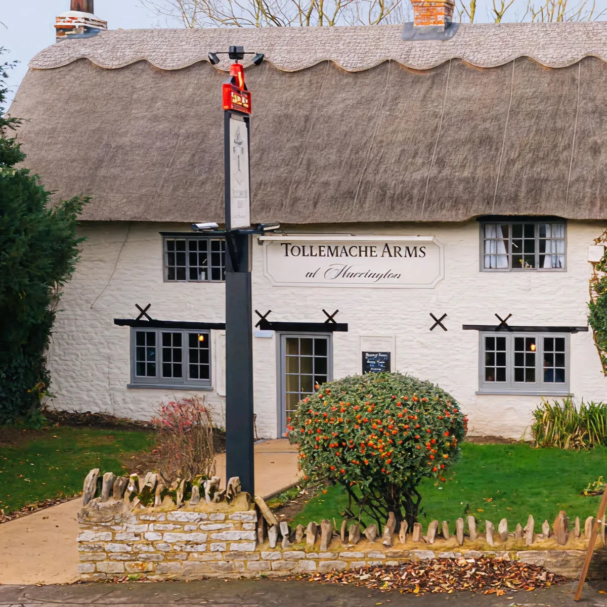 Thatched roof cottage named Tollemache Arms with a garden, stone fence, and a tall black post sign in front.