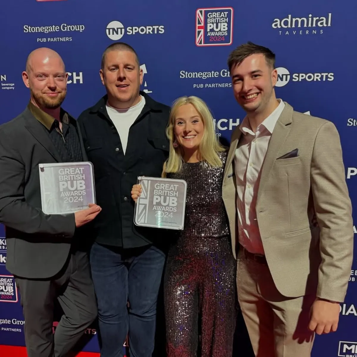 Four people dressed in formal attire smiling and holding Great British Pub Awards 2024 plaques in front of a blue sponsor backdrop.