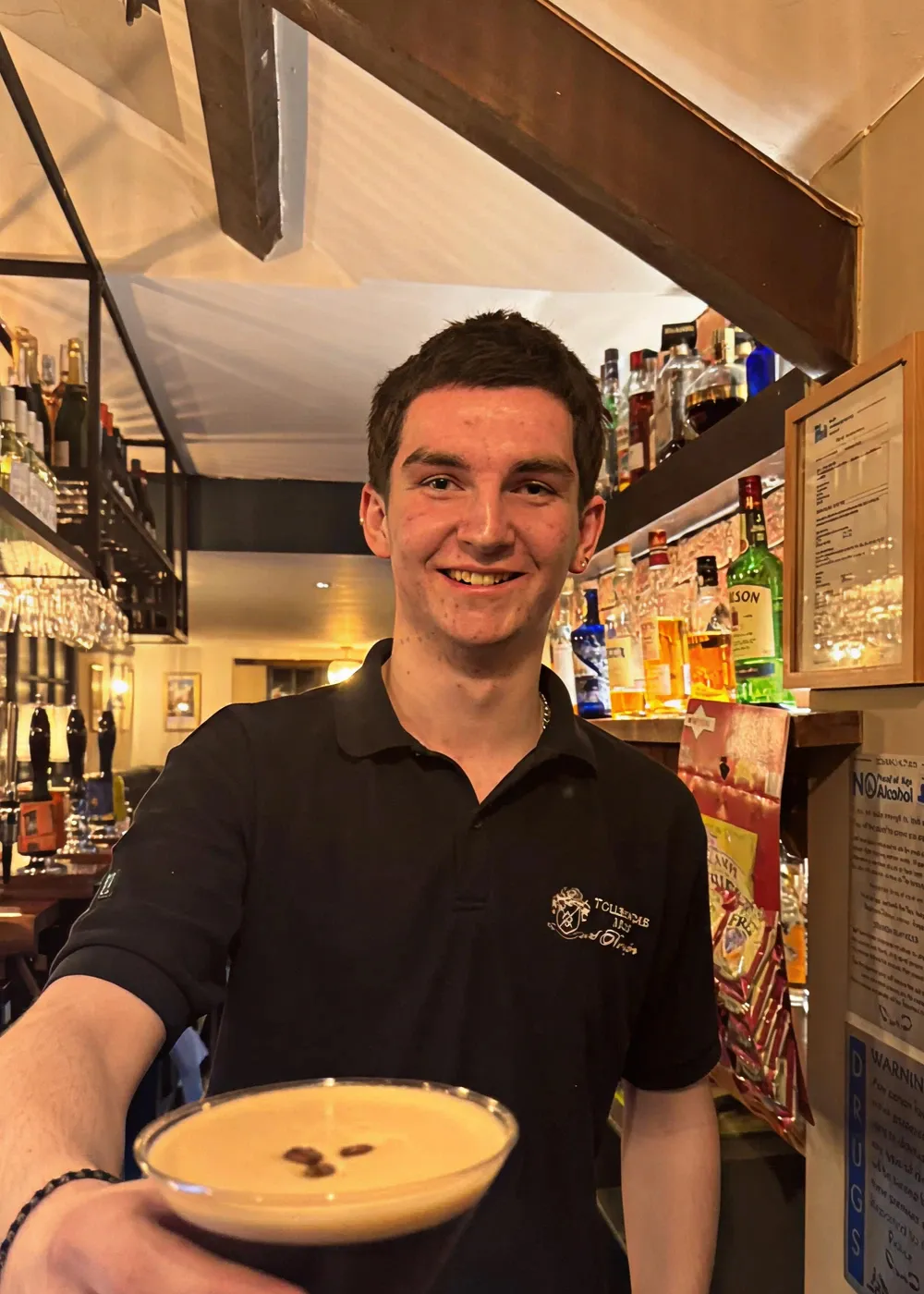 Smiling bartender in a black polo shirt holding out a cocktail glass with a creamy drink topped with coffee beans in a bar setting.