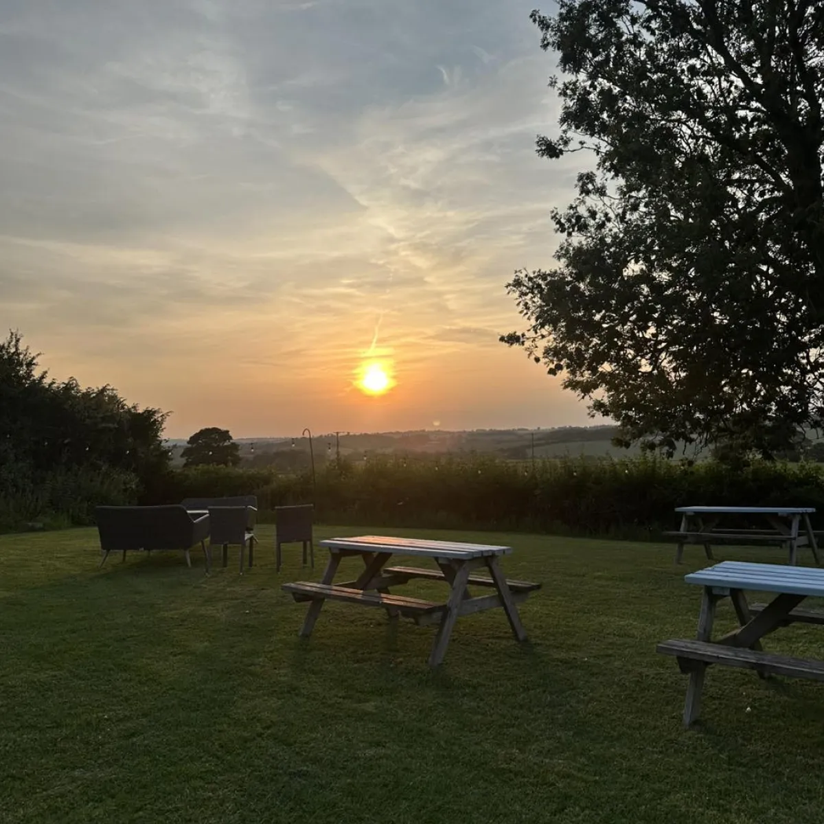 Sunset over a grassy outdoor area with wooden picnic tables, a seating area with chairs, and trees in the background.