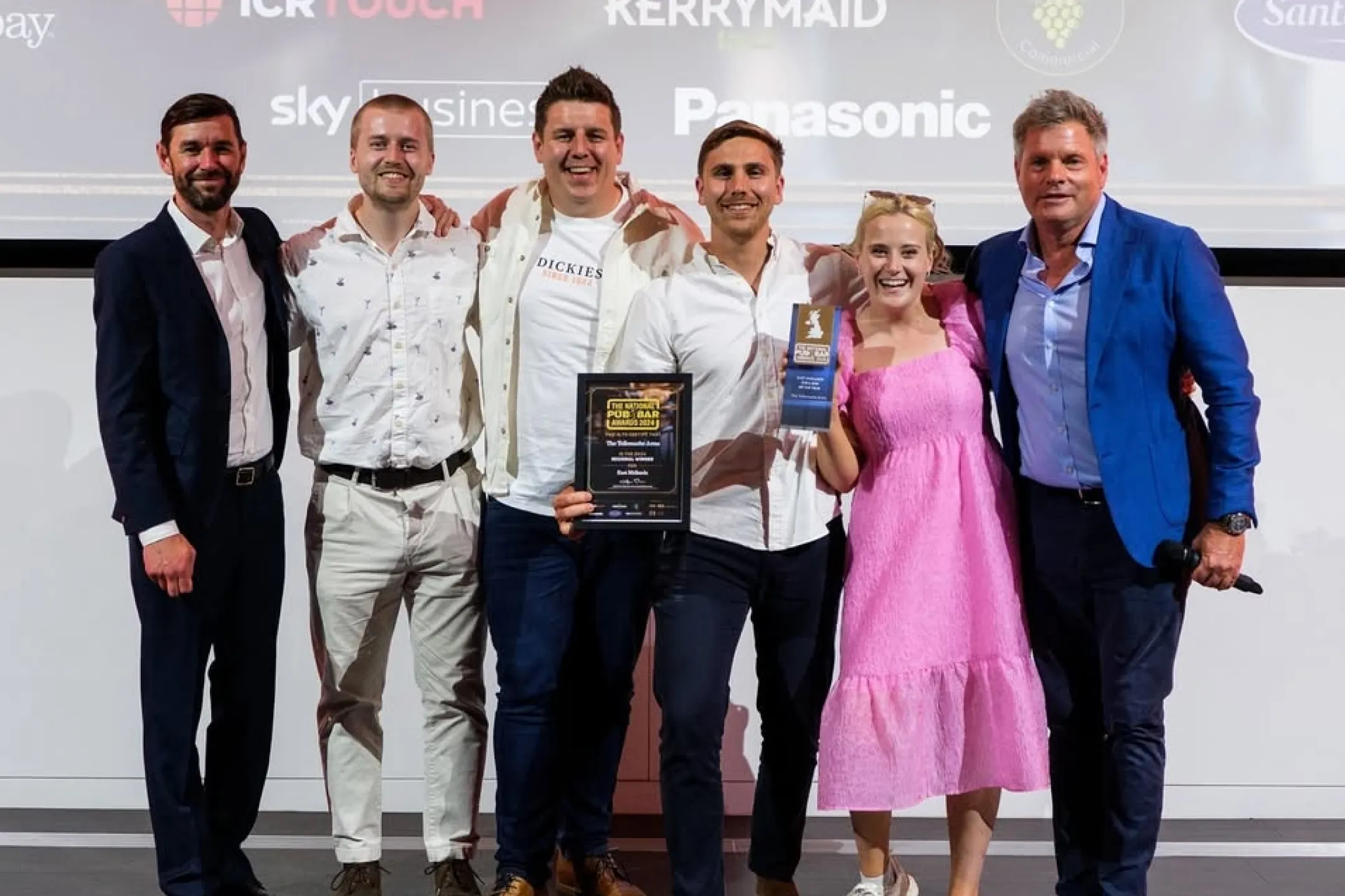 A group of six people standing together smiling, with one person holding a framed award and another holding a trophy at an event.