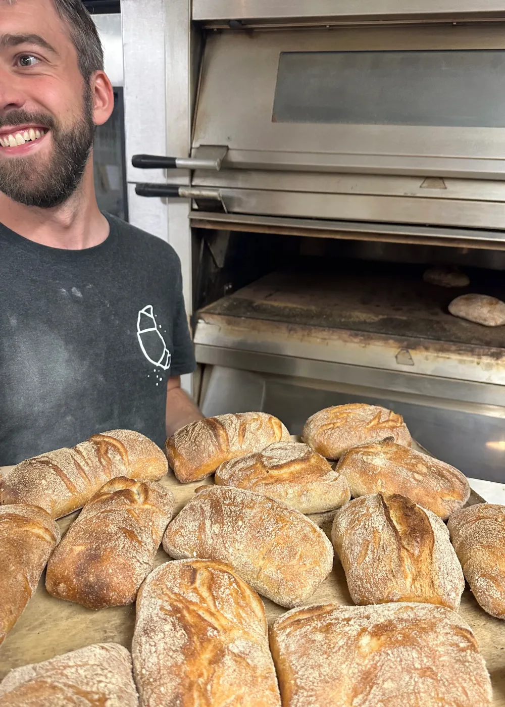 Smiling man holding a wooden tray with freshly baked crusty bread loaves in a bakery kitchen.