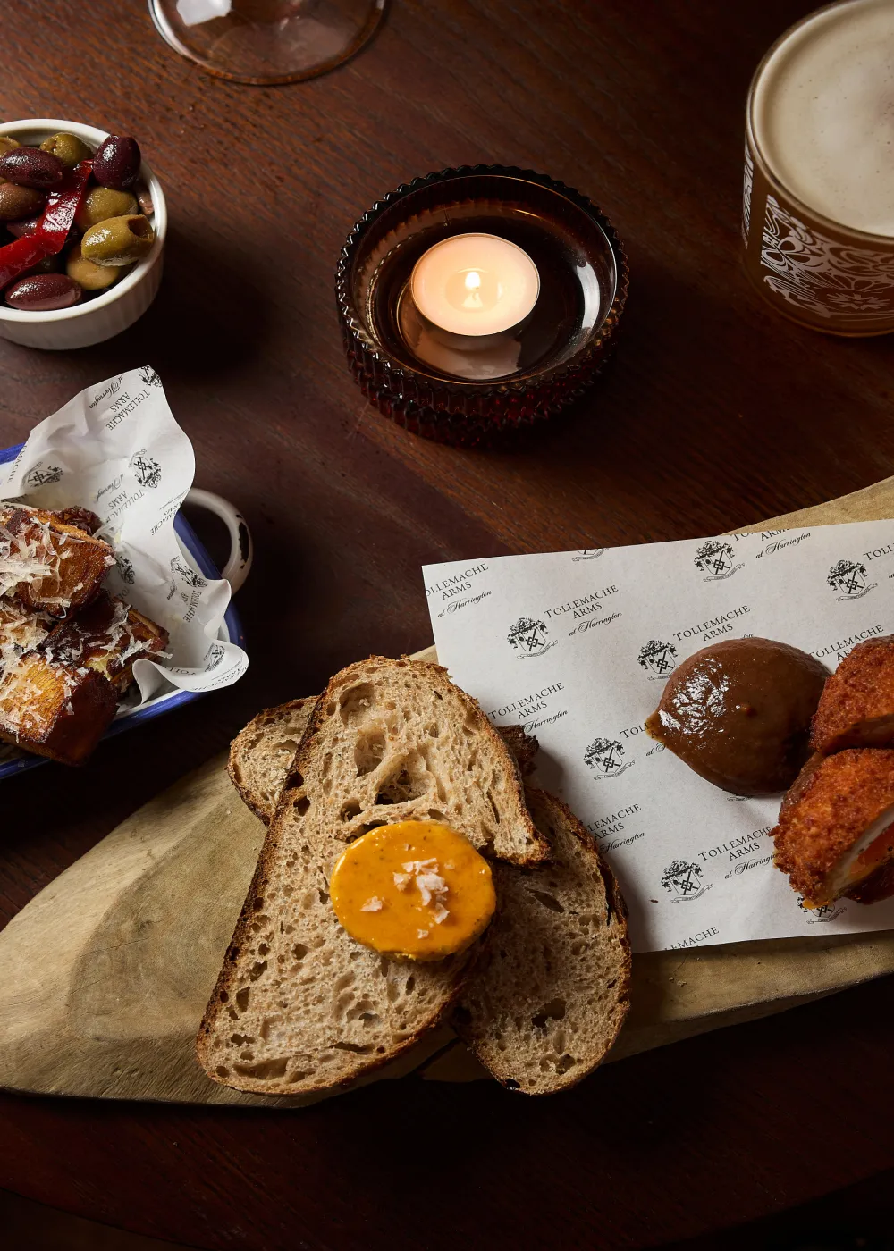 Wooden table with slices of bread topped with a yellow spread, a bowl of mixed olives, fried food on branded paper, and a lit candle in a small holder.