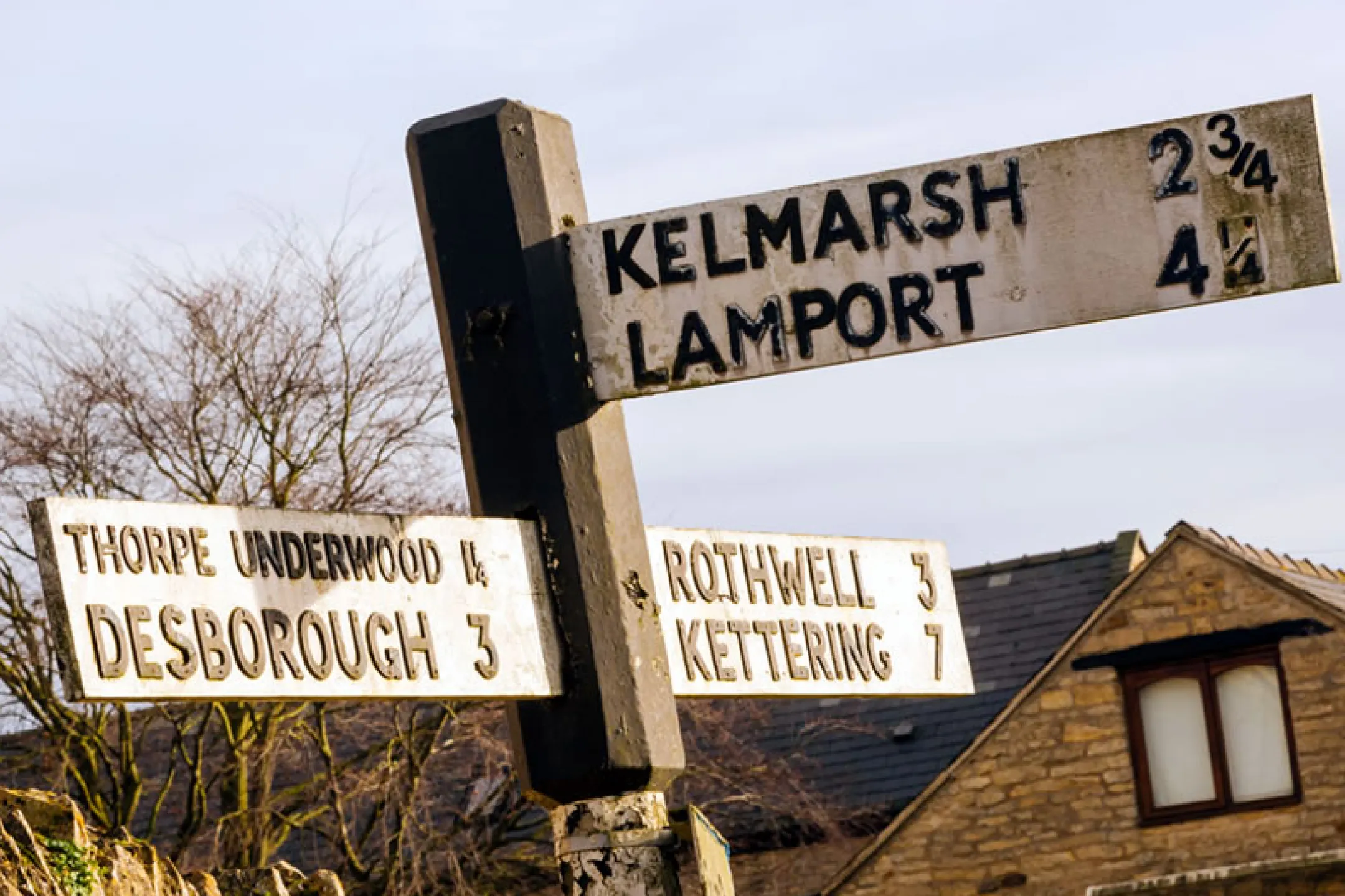 Old-style directional road signs pointing to Kelmarsh, Lamport, Thorpe Underwood, Desborough, Rothwell, and Kettering with distances in miles.