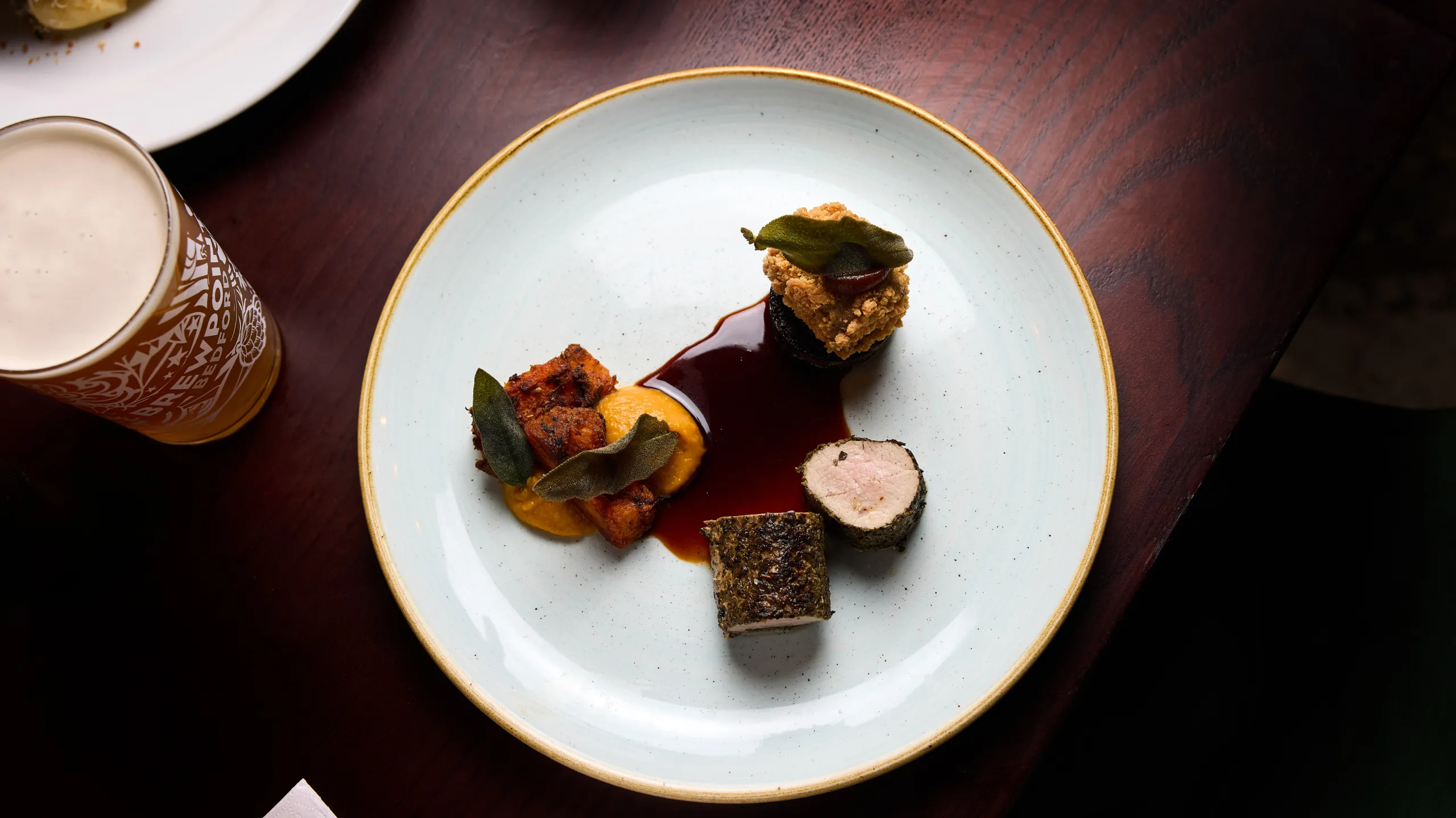 Gourmet dish on a white plate with three small pieces of meat, two dollops of sauce, herbs on top, and a glass of beer on a dark wooden table.