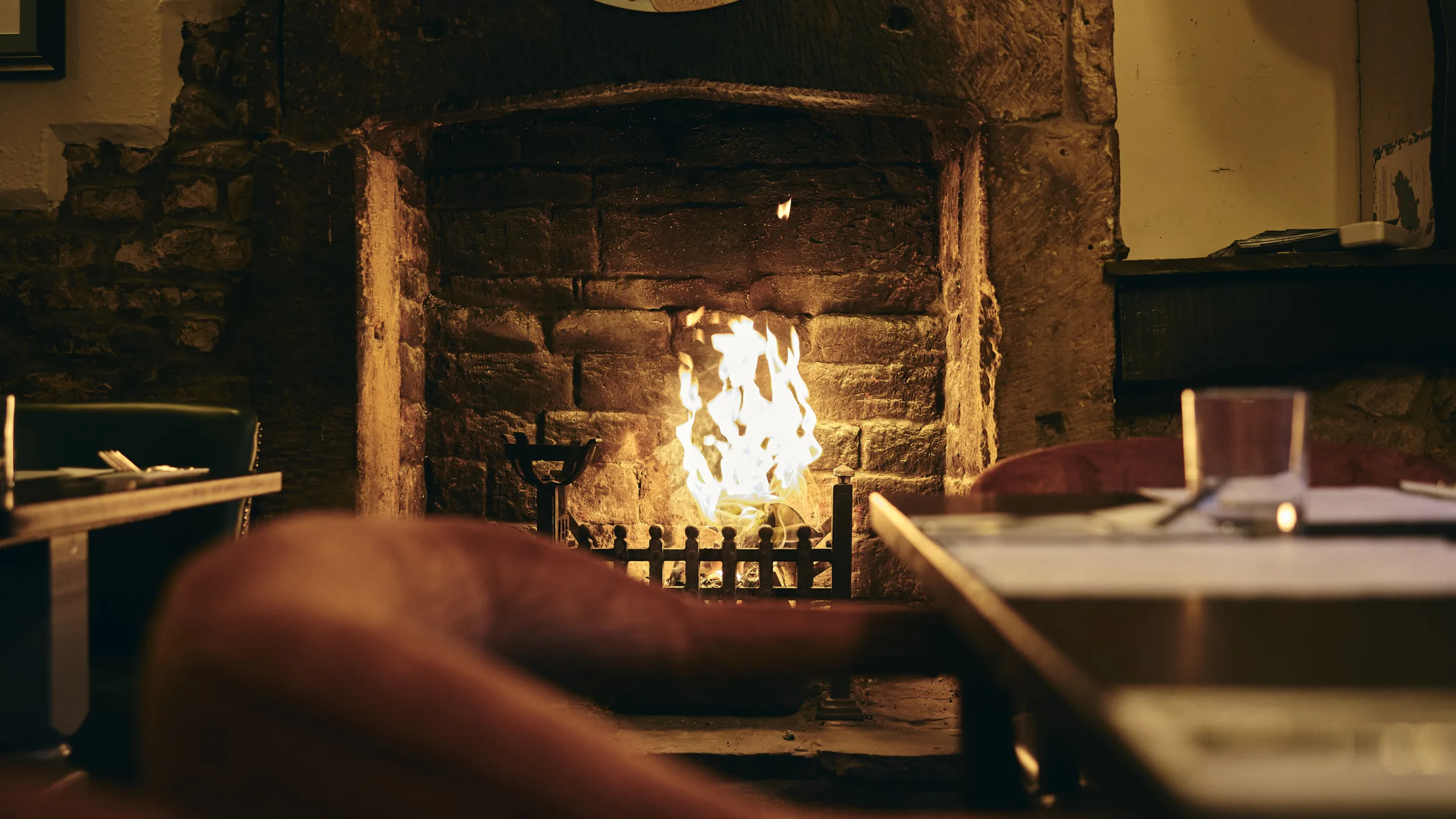 Cozy stone fireplace with a bright fire burning, surrounded by tables and chairs in a warm room.