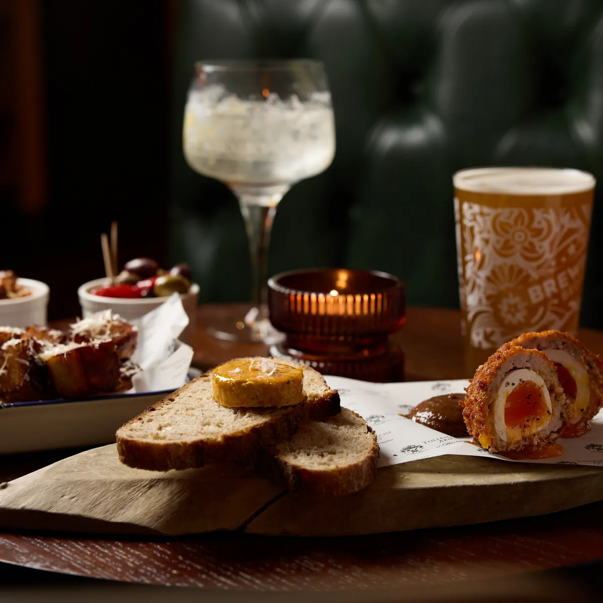 Wooden board with slices of bread topped with butter, Scotch eggs, a dish of pickled olives and cherry tomatoes, and artistic drinks on a dark restaurant table.
