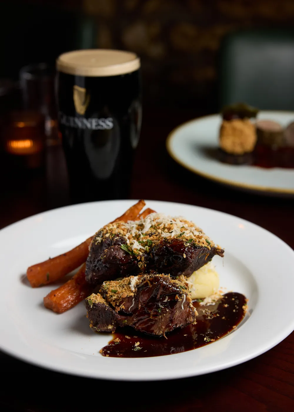 Plate with braised beef topped with grated cheese and herbs, served with mashed potatoes, glazed carrots, and dark sauce, with a pint of Guinness in the background.