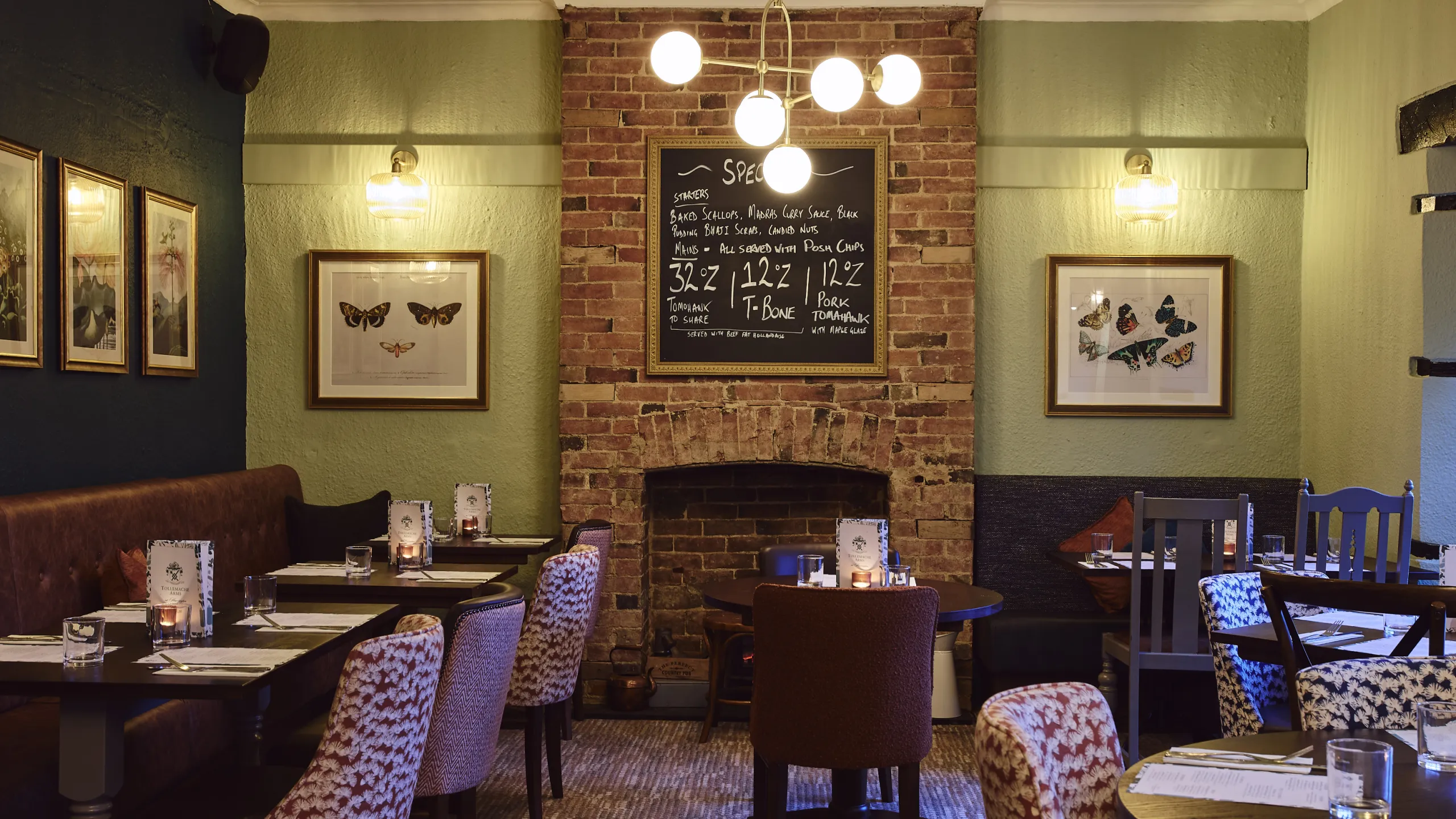 Cozy restaurant interior with a brick fireplace, a chalkboard menu, framed butterfly artwork, and tables set with menus and candles.