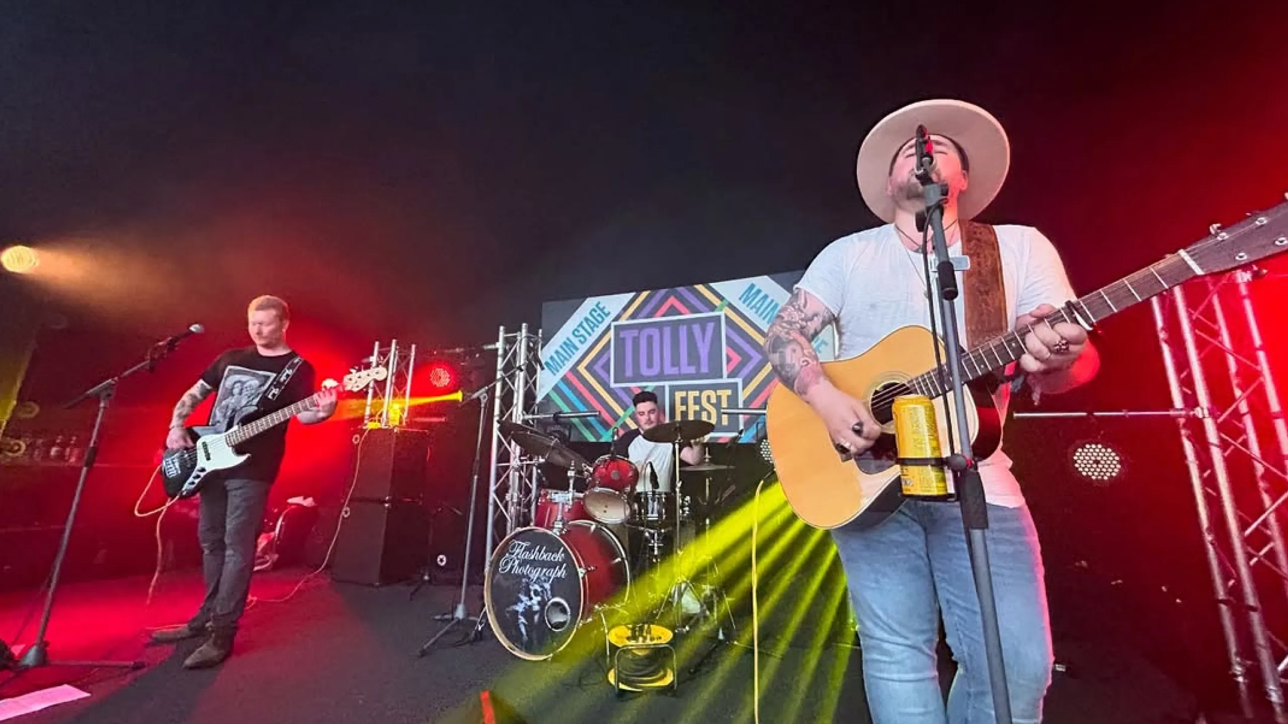 Three-member band performing live on stage at Tolly Fest with guitar, bass, and drums under colorful stage lights.
