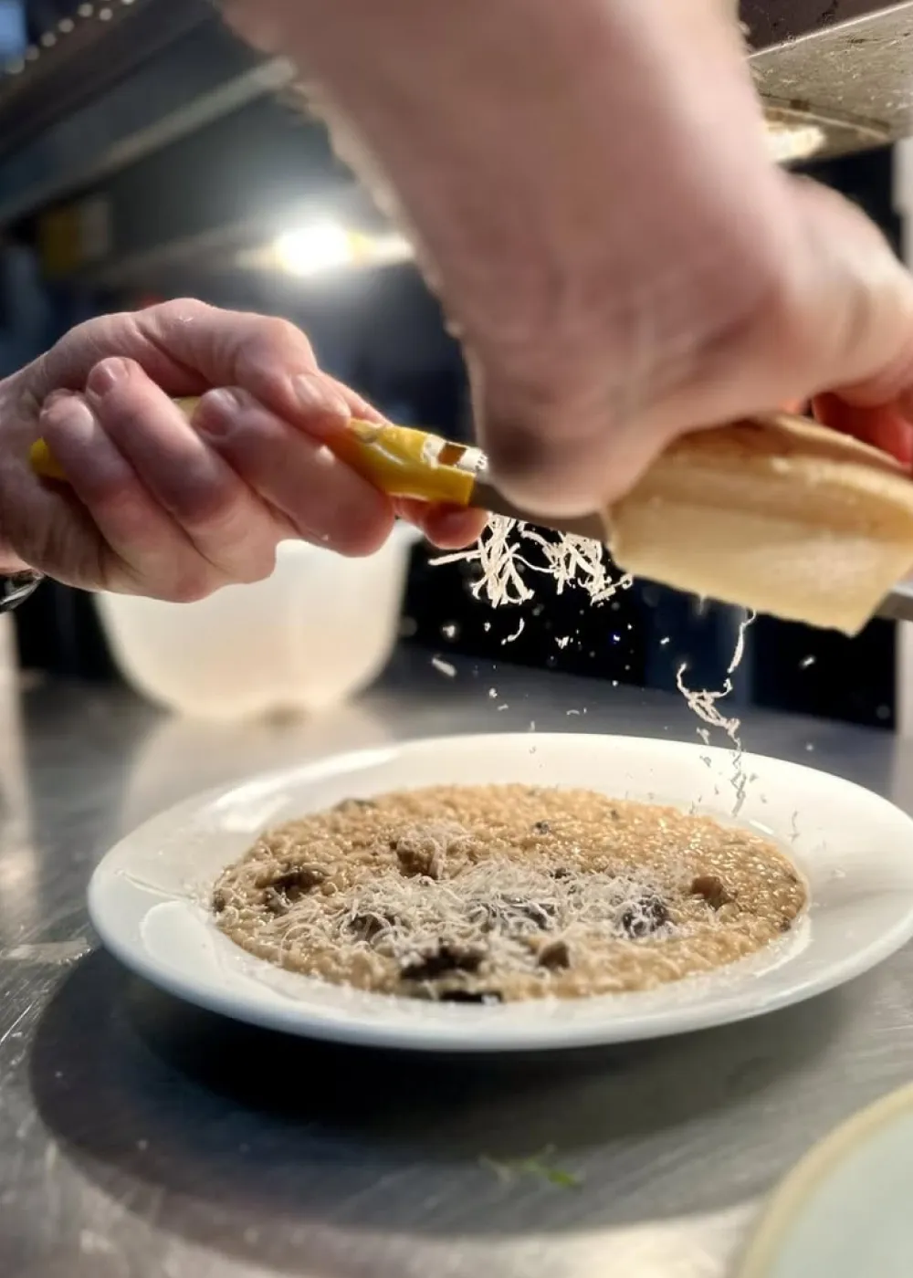 Close-up of hands grating cheese over a plate of risotto on a stainless steel surface.
