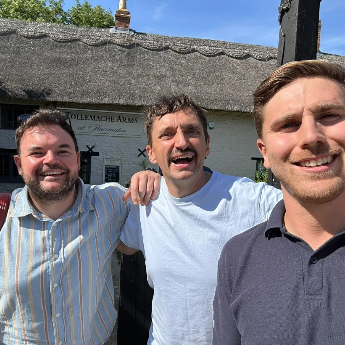 Three smiling men standing close together outdoors in front of a building with a sign reading TOLLEMACHE ARMS at Harrington.