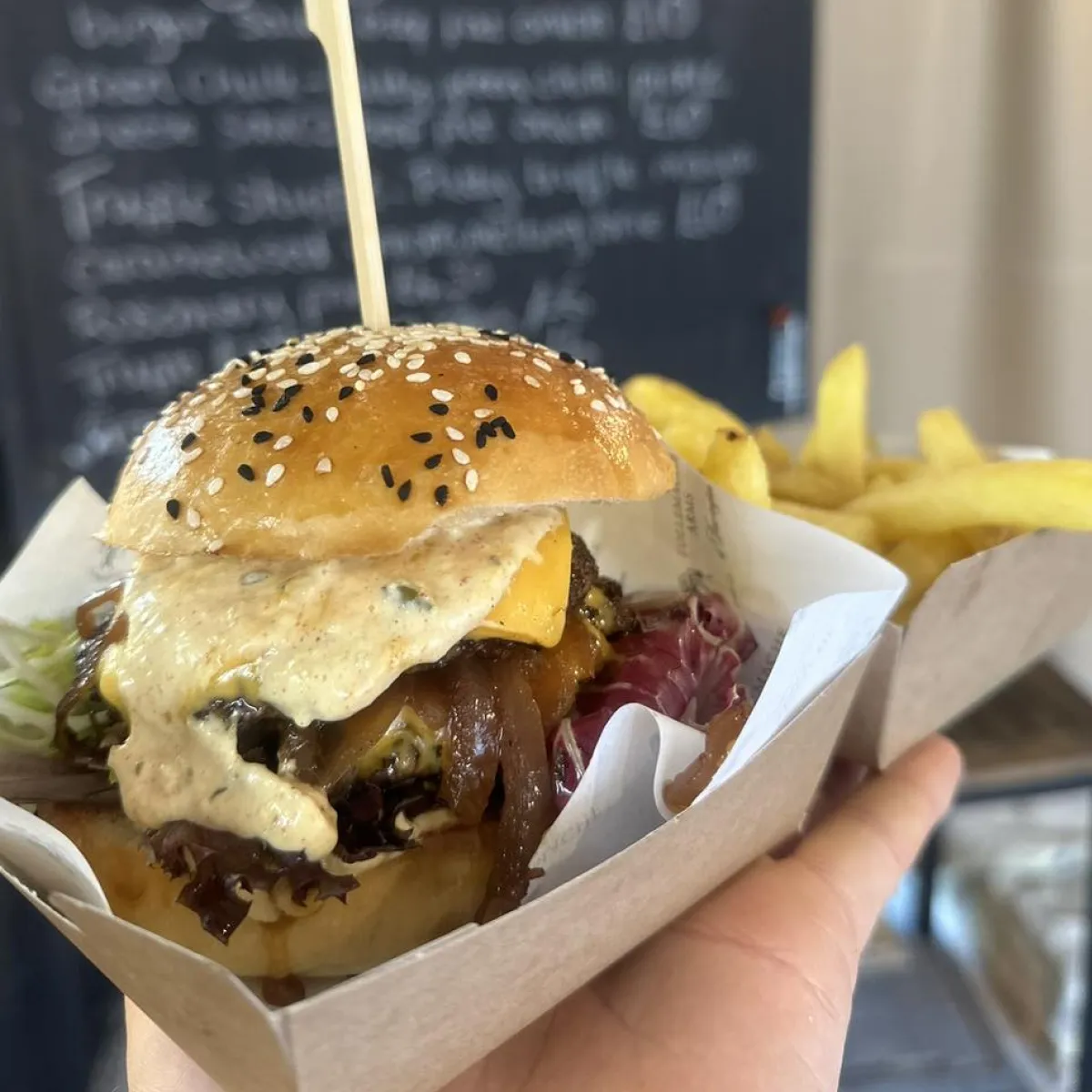 Hand holding a cheeseburger with sesame seed bun, lettuce, caramelized onions, cheese, sauce, and a side of French fries in a paper tray.