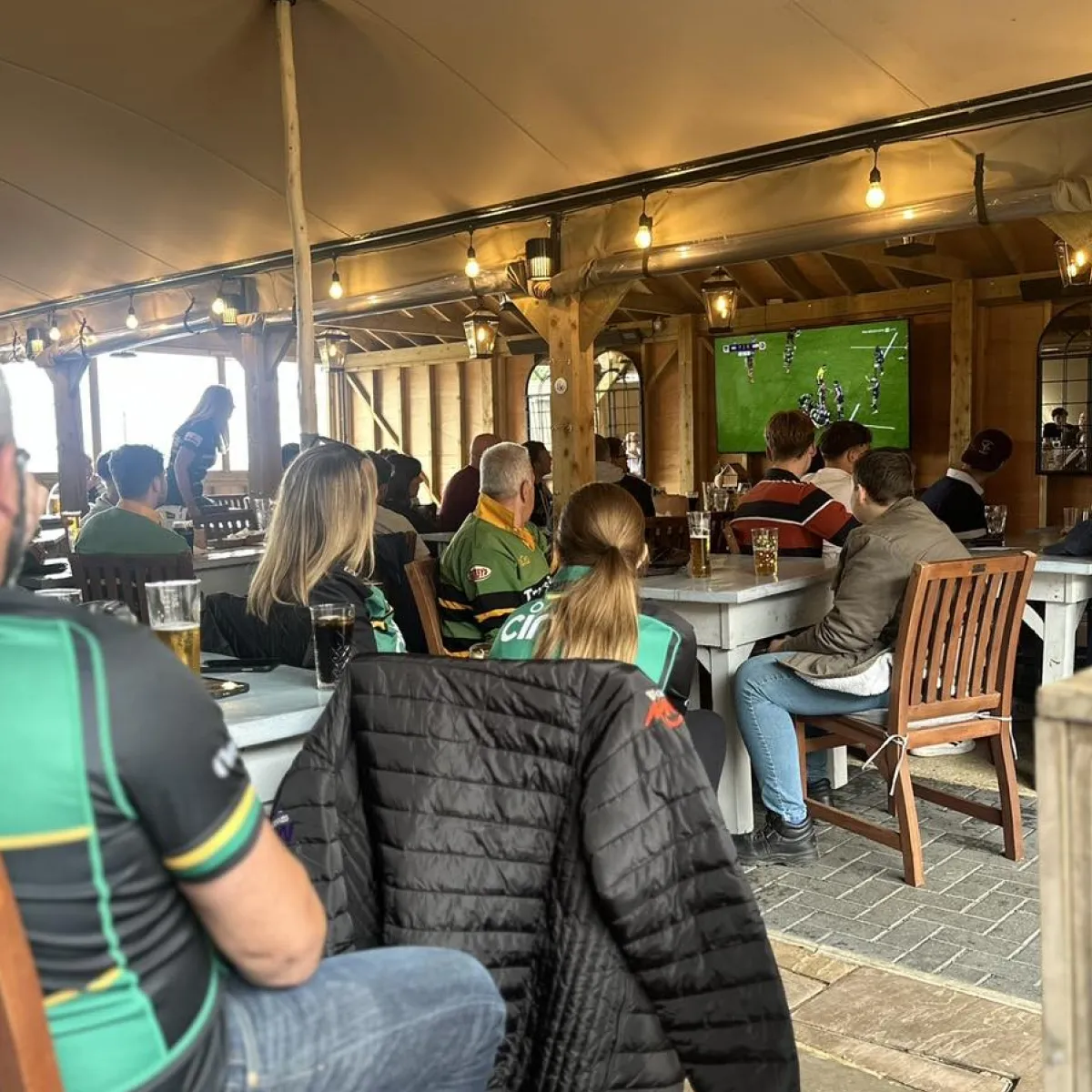 Group of people sitting in a wooden indoor space watching a rugby match on a mounted TV screen.