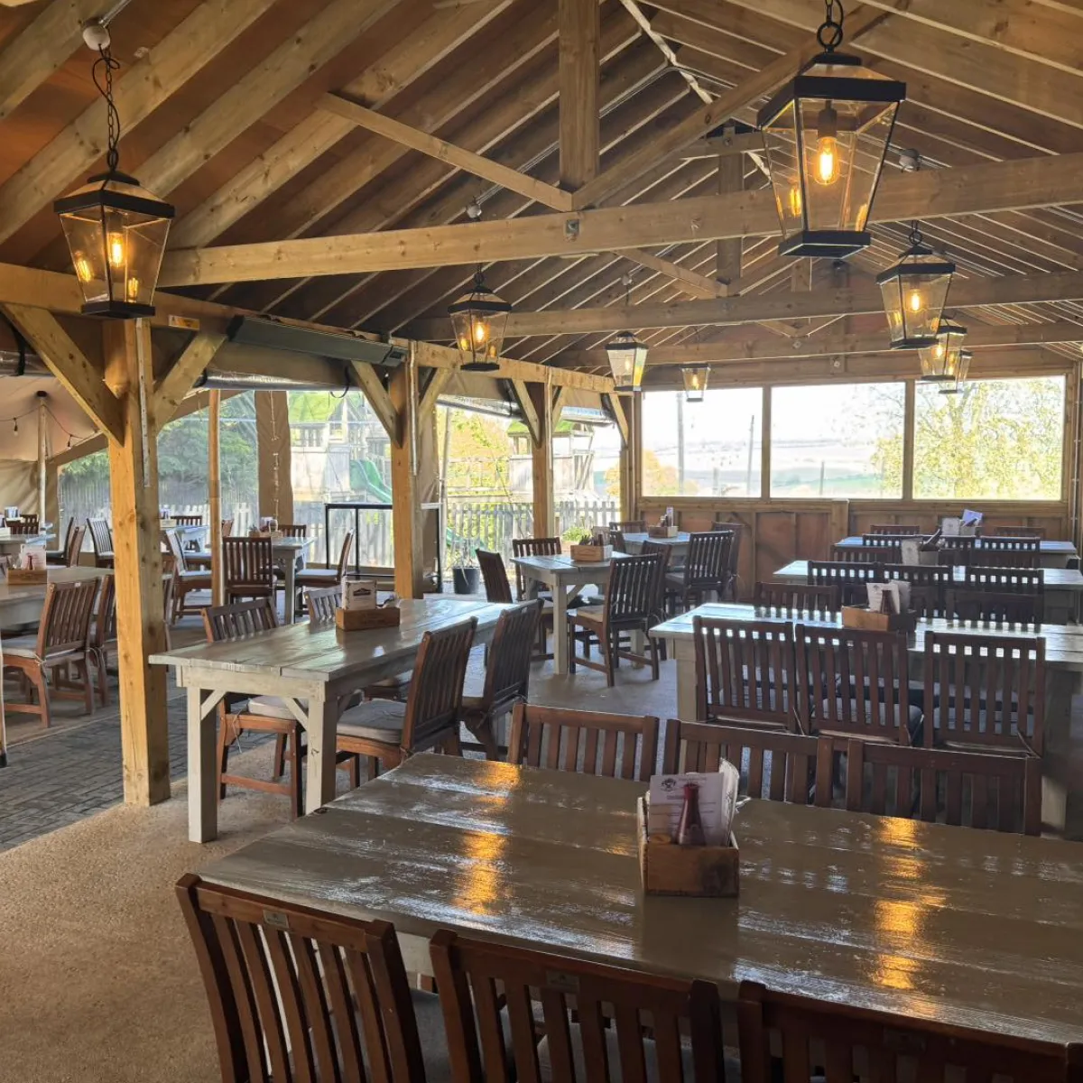 Indoor rustic dining area with wooden tables and chairs under exposed wooden beams and hanging lantern lights.