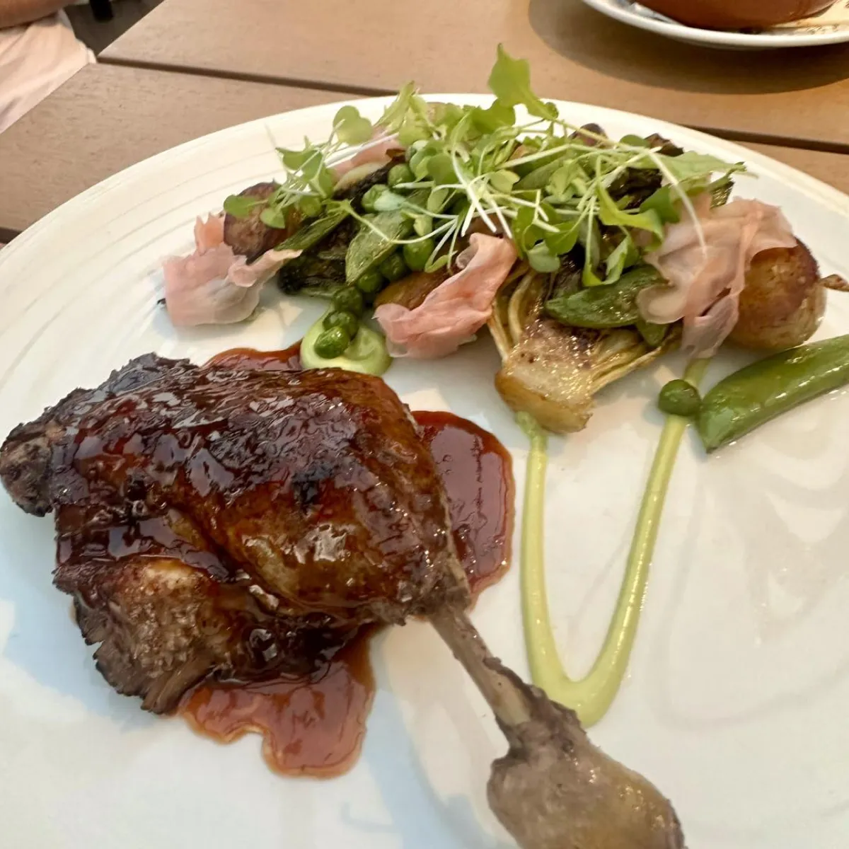Plate with roasted duck leg glazed in sauce, accompanied by grilled vegetables, peas, microgreens, and thinly sliced pickled ginger.