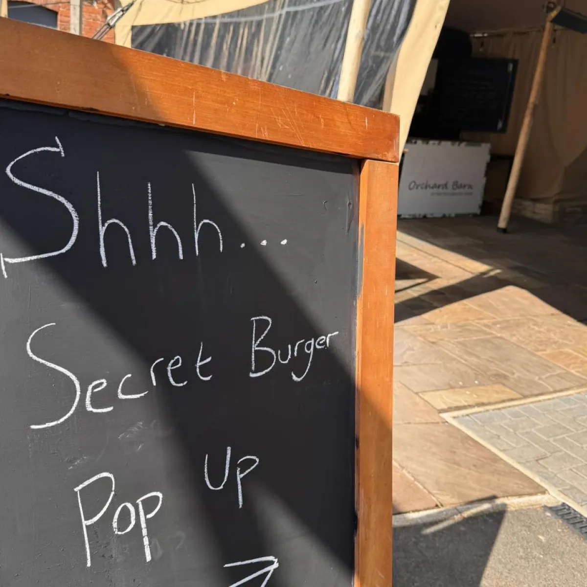 Chalkboard sign with wooden frame that says 'Shhh... Secret Burger Pop Up' with an arrow pointing right in front of a building entrance.