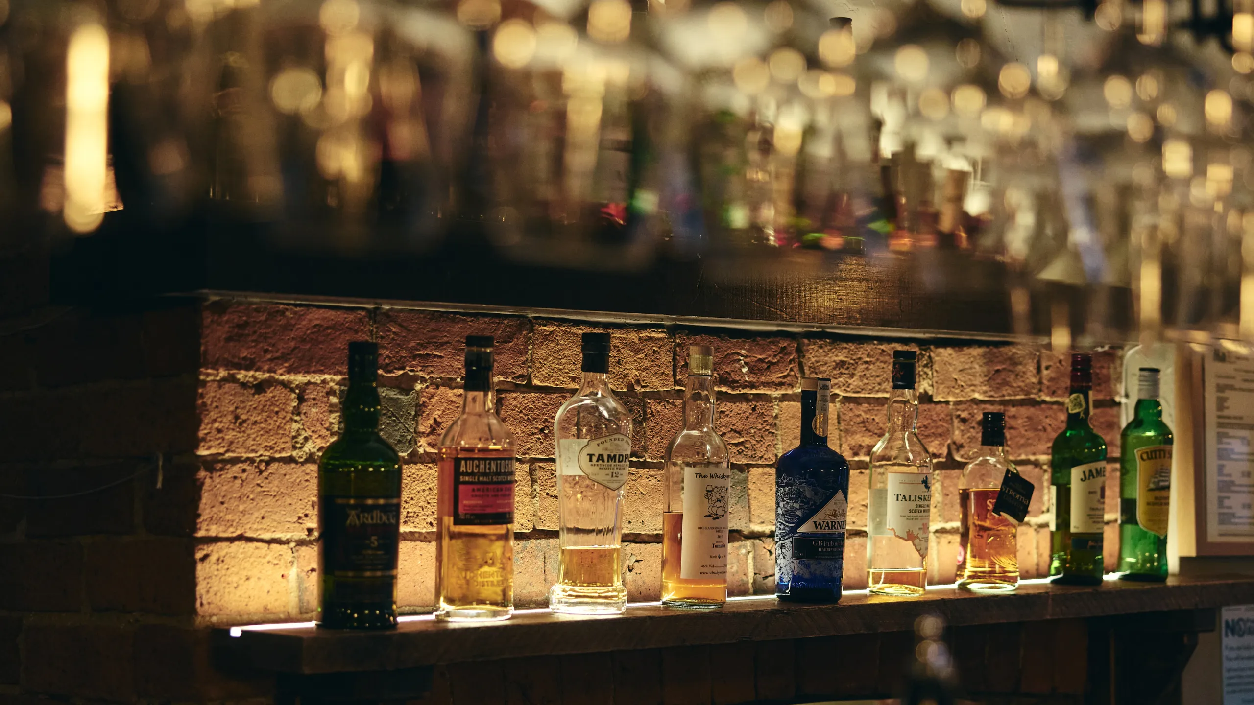 Backlit bar shelf with various bottles of whiskey and liquor against a brick wall.