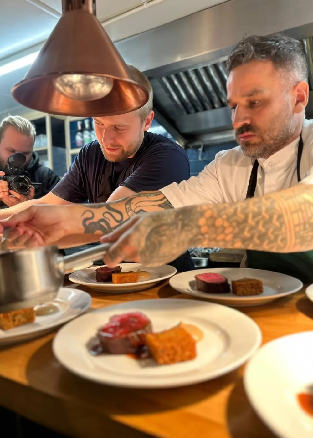 Two tattooed chefs carefully plating gourmet dishes in a professional kitchen while a person films them.