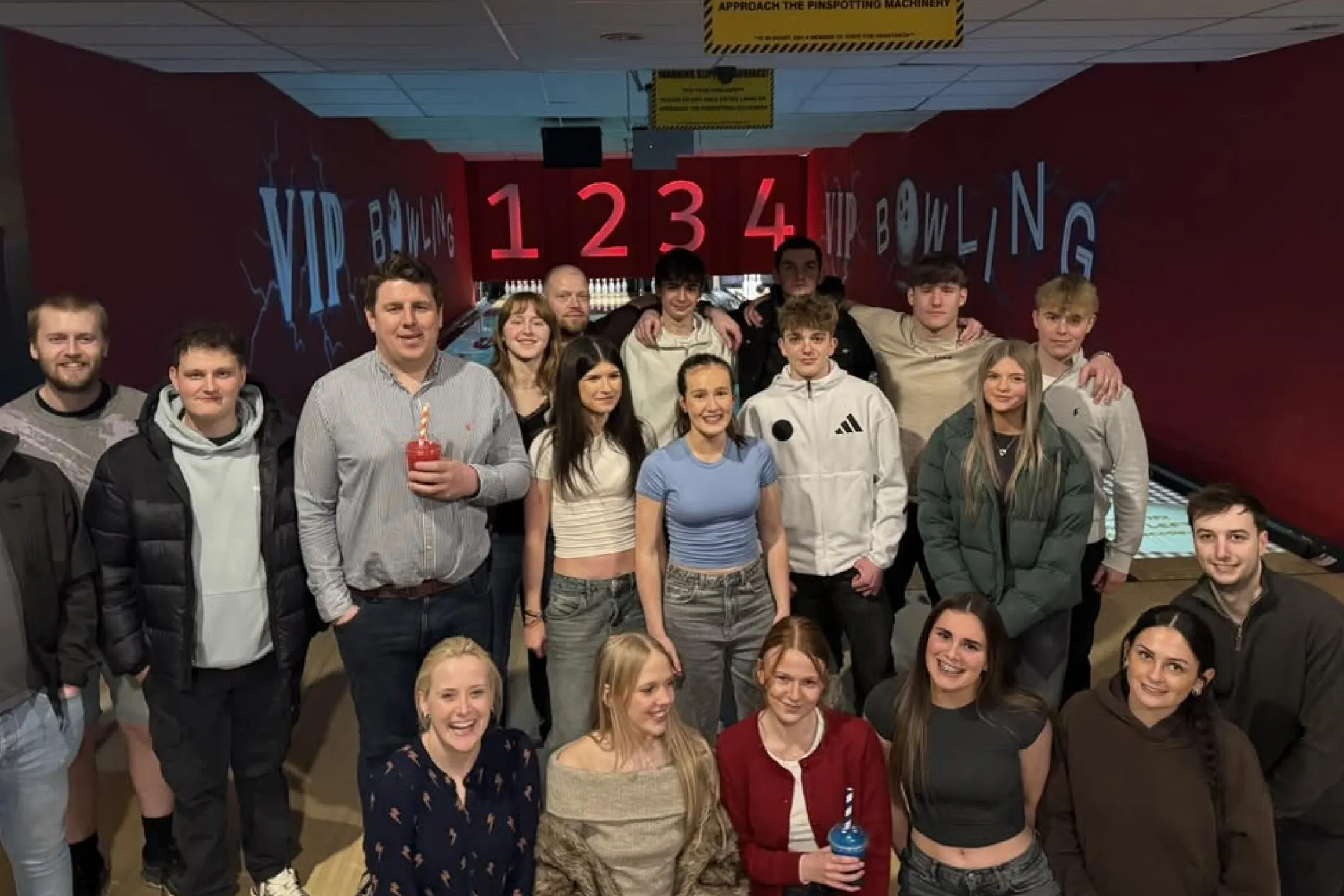 Group of young adults smiling and posing together in a bowling alley with lanes and pins visible in the background.