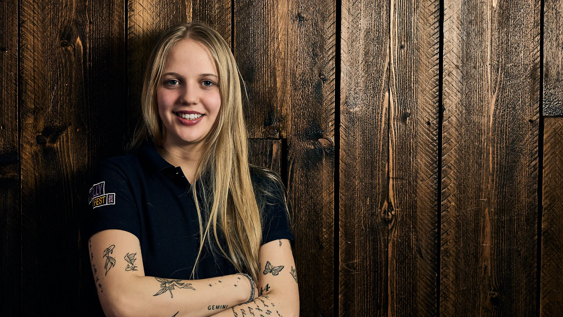 Smiling blonde woman with tattoos on both arms standing with arms crossed in front of a dark wooden wall.