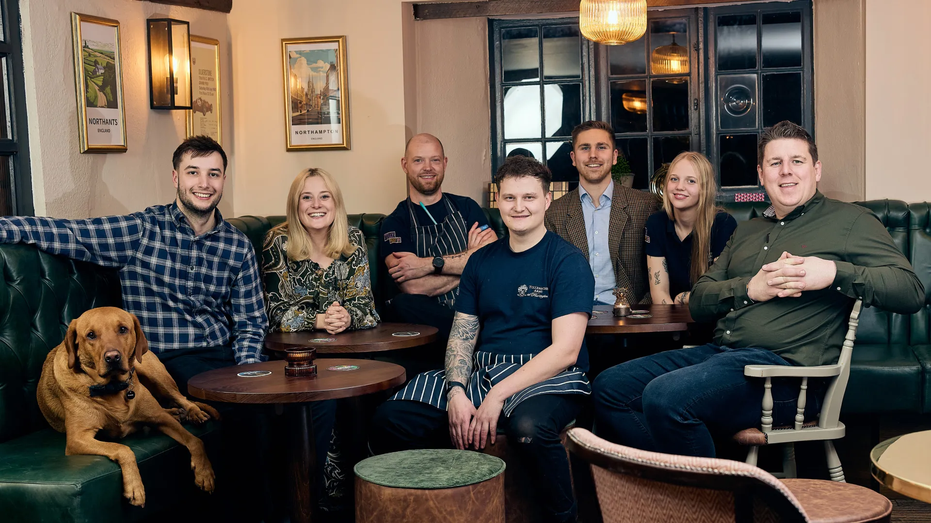 Group of six people and a brown dog seated around wooden tables in a cozy pub setting with framed pictures and pendant lights.