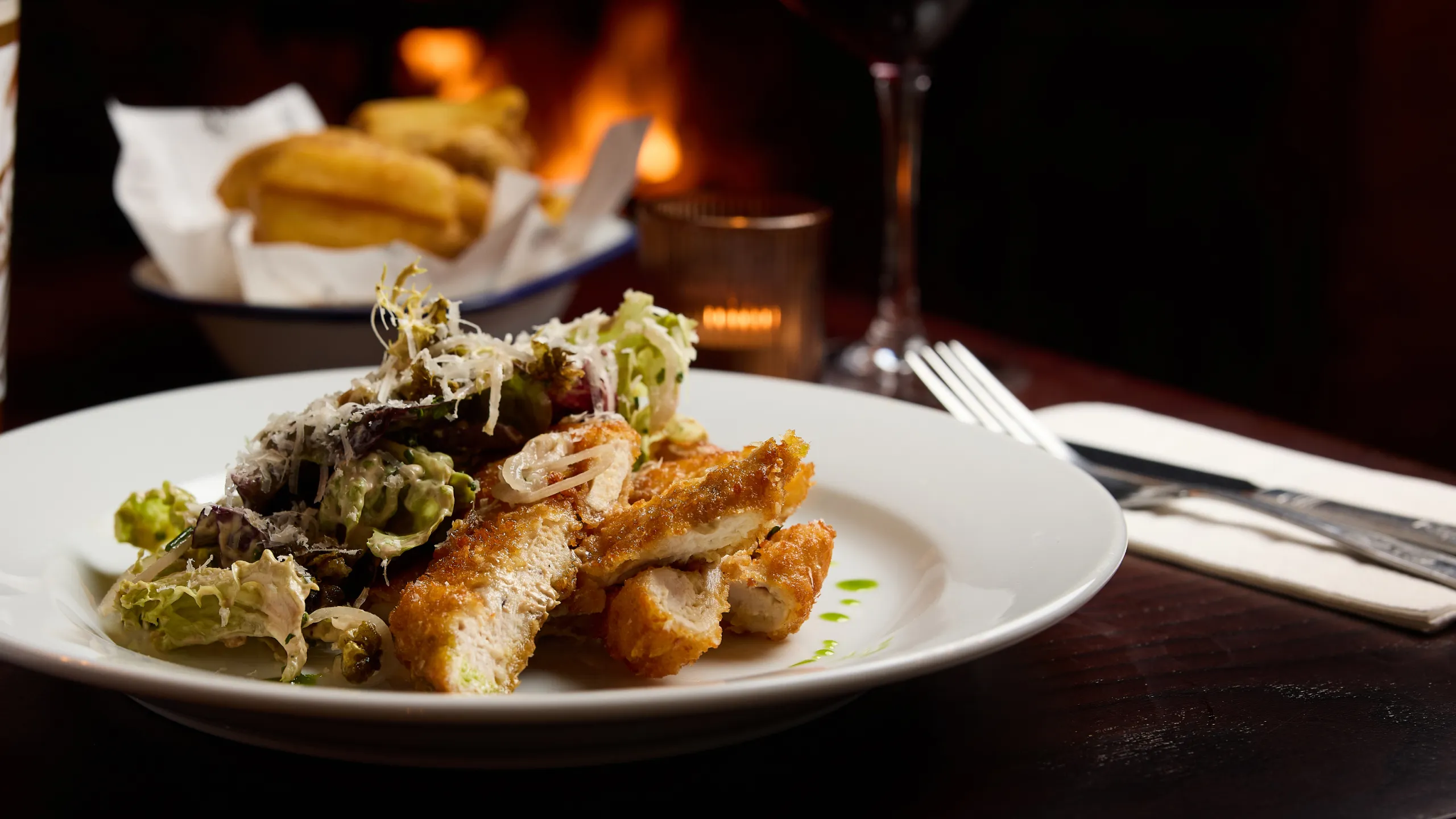 Plate of crispy breaded chicken strips served with a creamy salad and grated cheese, with bread rolls and a lit candle in the background.