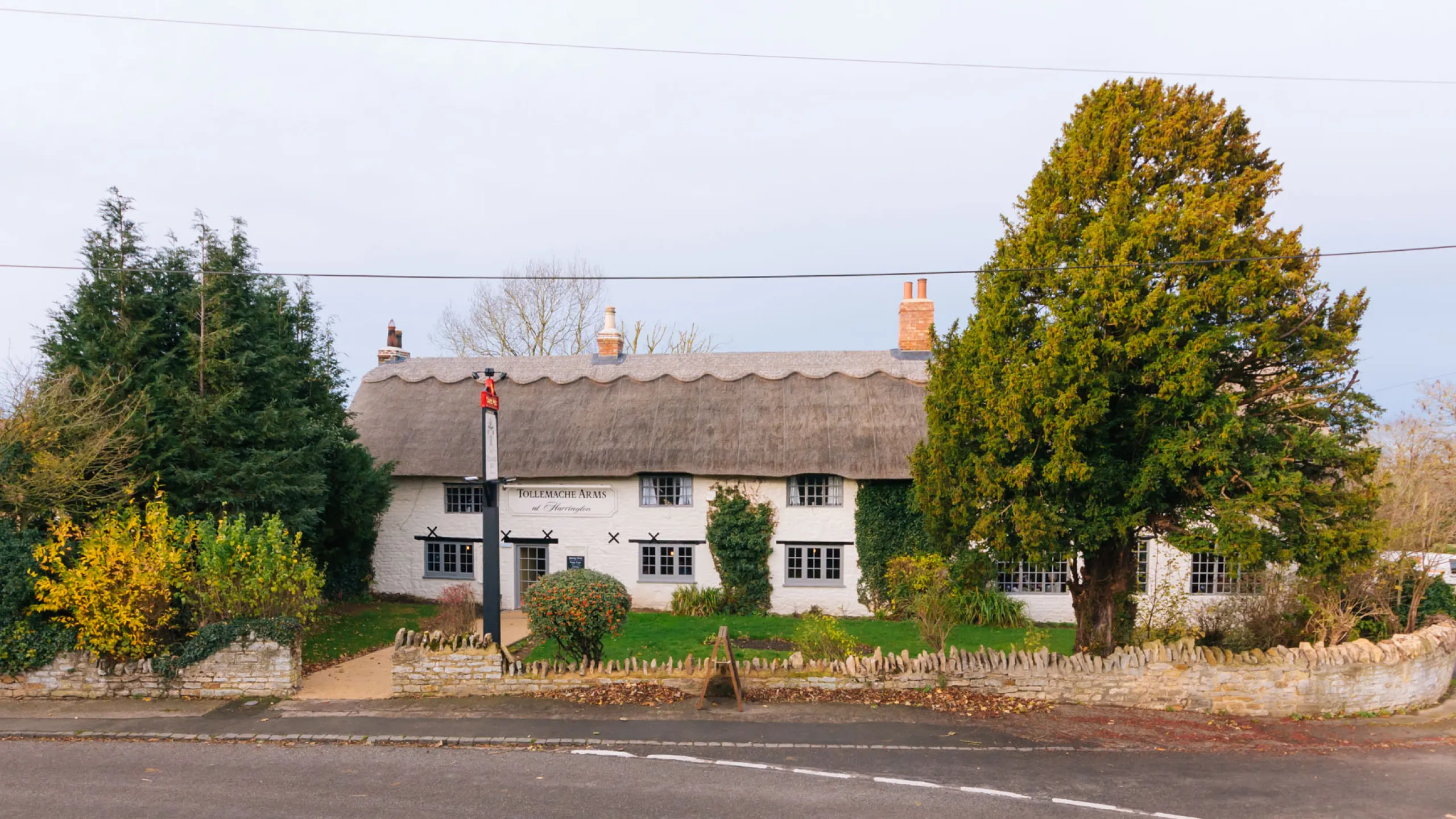Thatched roof white building labeled Tollemache Arms surrounded by large trees and a stone fence.