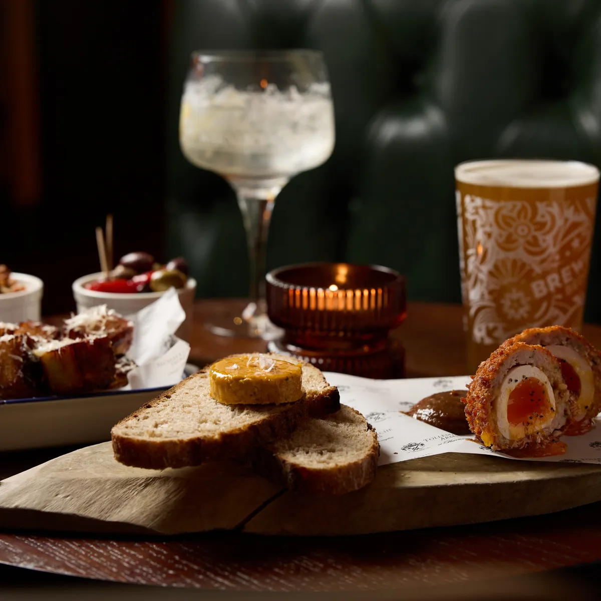 Wooden board with slices of bread topped with butter, Scotch eggs cut in half with sauce, small bowls of olives and snacks, a lit candle, a cocktail glass, and a glass of beer on a dark wooden table.