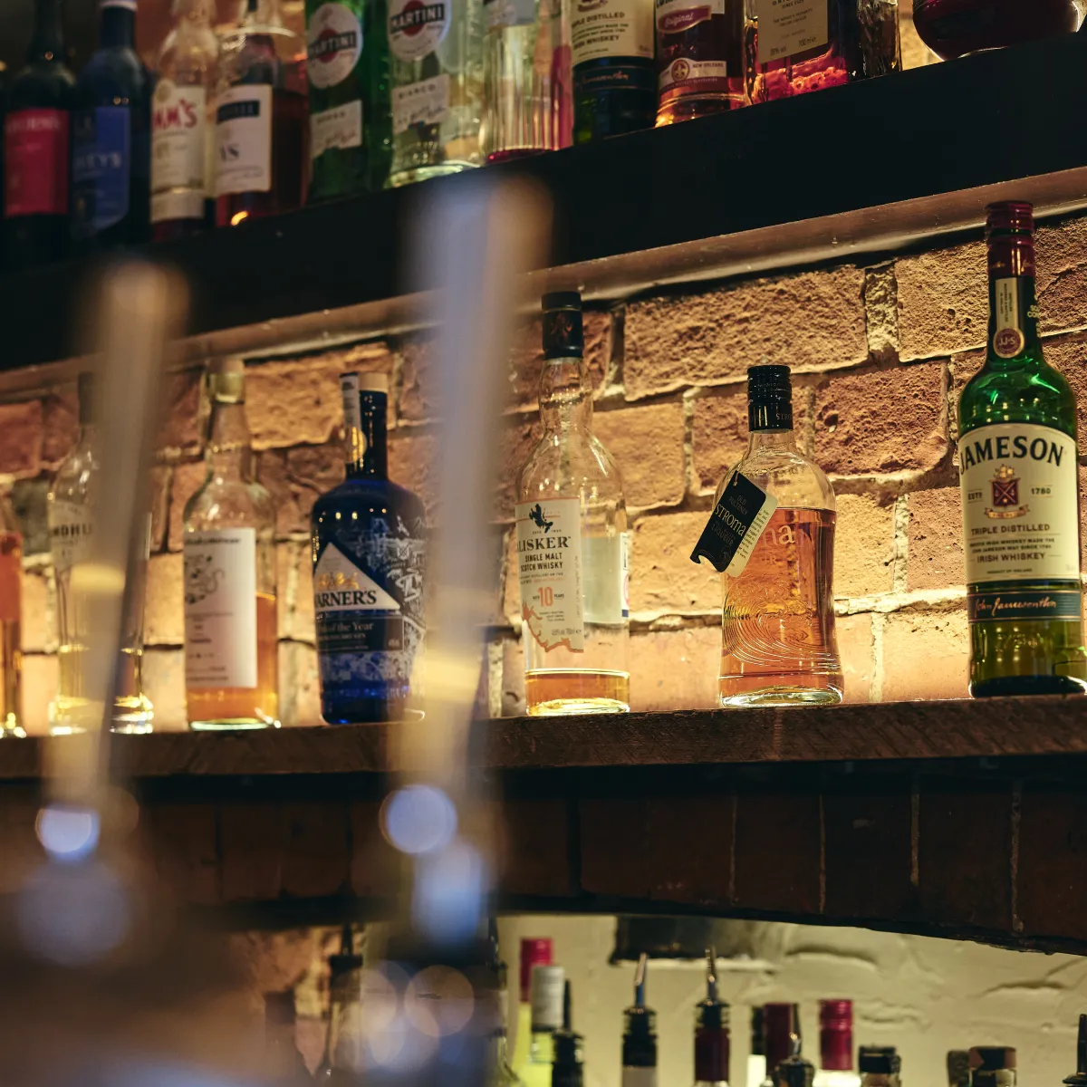 Various liquor bottles including Jameson Irish whiskey displayed on wooden shelves against a brick wall background in a dimly lit bar.