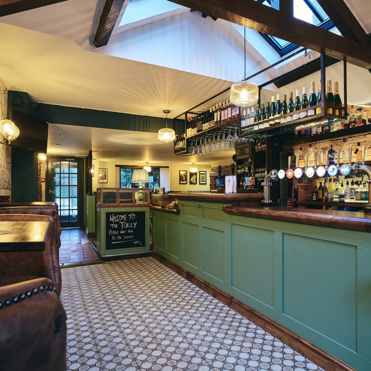 Interior of a cozy pub with green paneled bar, hanging wine bottles, beer taps, pendant lights, and cushioned booths.
