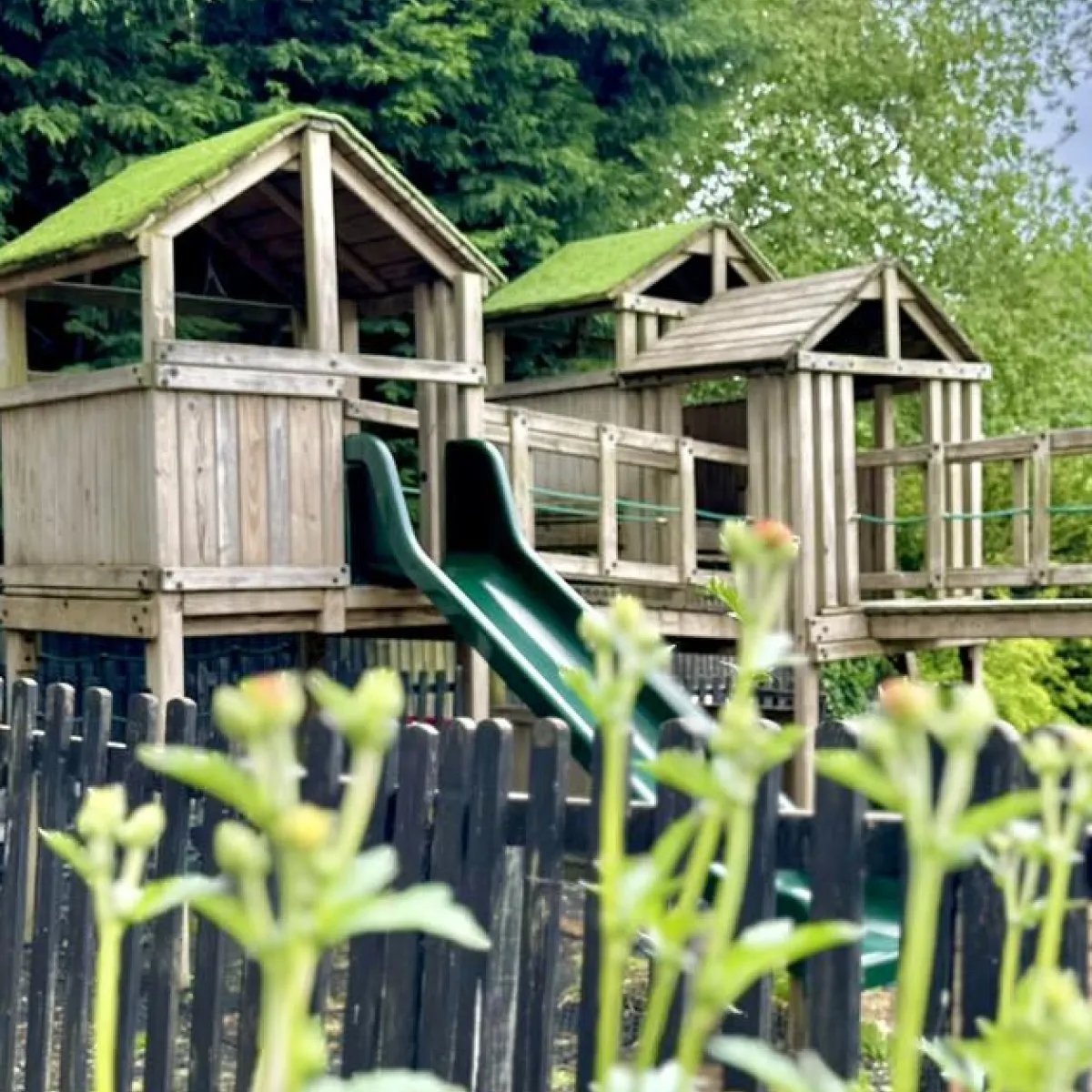 Wooden playground structure with green slides and moss-covered roofs surrounded by greenery.