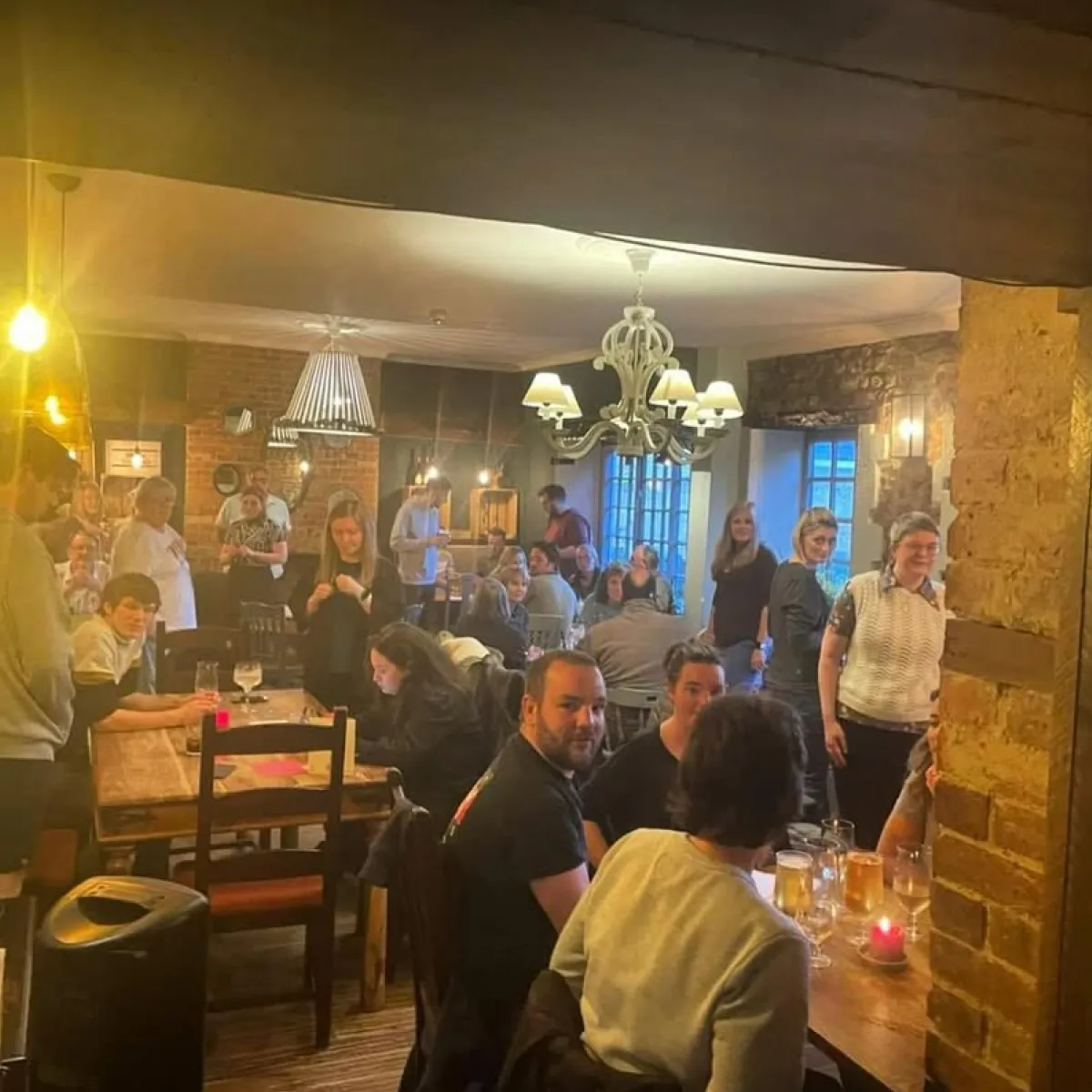 Group of people socializing and dining in a warmly lit rustic restaurant with wooden tables and brick walls.