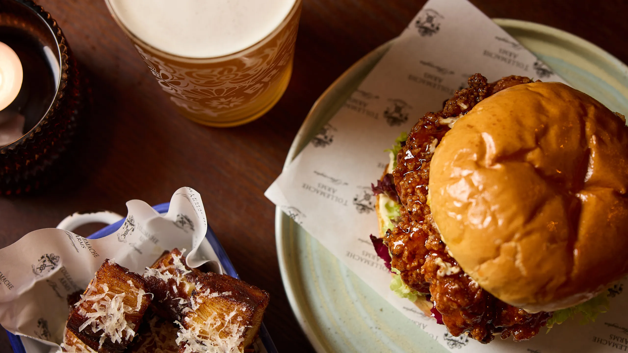 Spicy fried chicken sandwich on a plate with crispy potato cubes topped with grated cheese and a glass of beer on a wooden table.