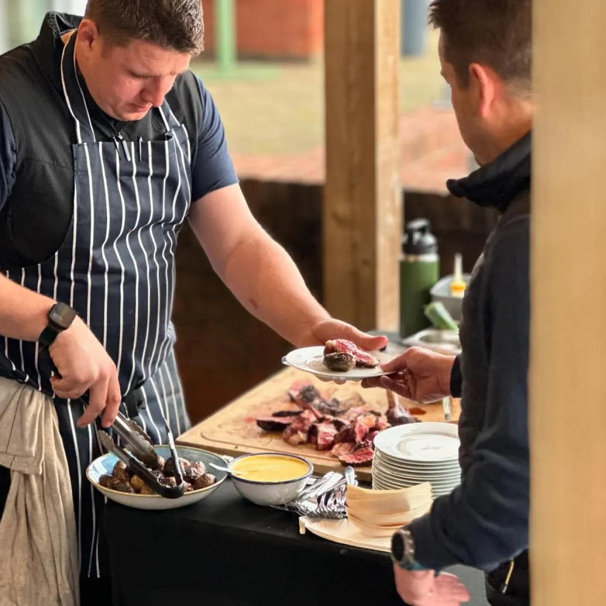 Chef in striped apron serving sliced cooked meat onto a plate for a guest at a buffet table with bowls of sauce and plates.