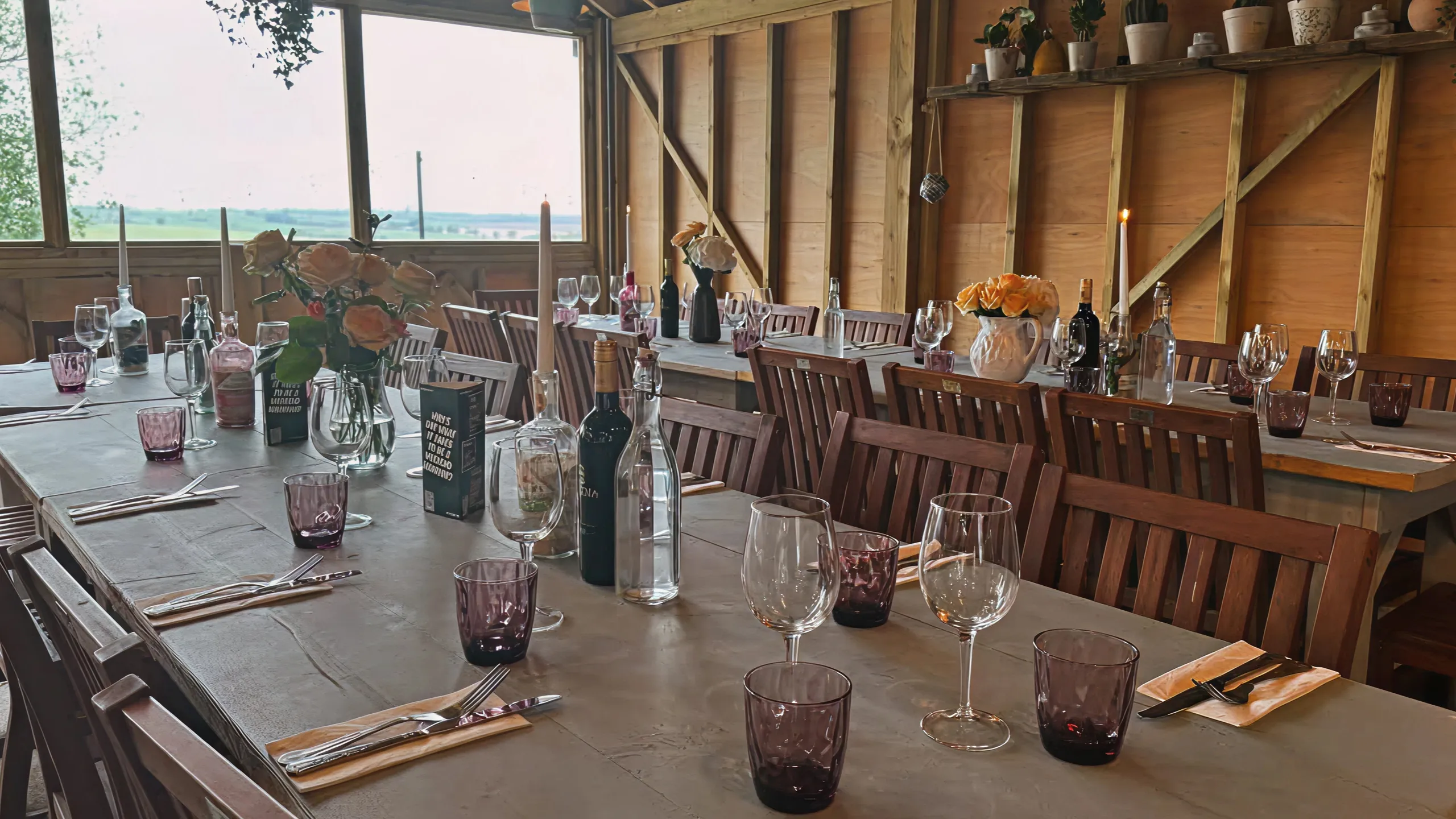 Rustic dining room with long wooden tables set with wine glasses, purple tumblers, flowers, and candles.