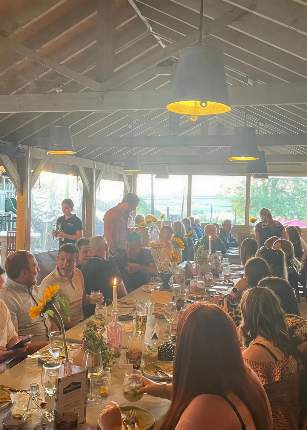 Large group of people dining together at a long table decorated with sunflowers and candles in a rustic indoor setting with wooden beams and pendant lights.