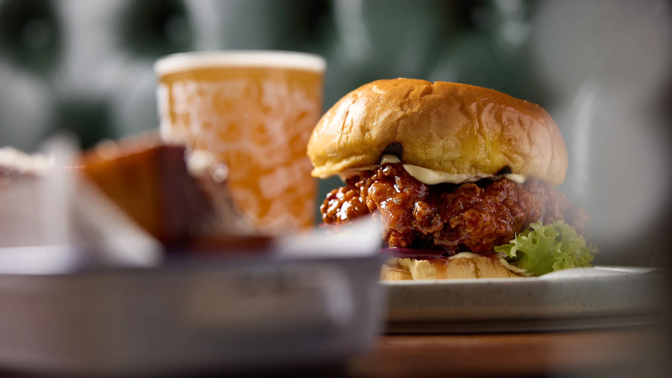 Close-up of a fried chicken sandwich with lettuce and mayonnaise on a bun, with a glass of beer blurred in the background.