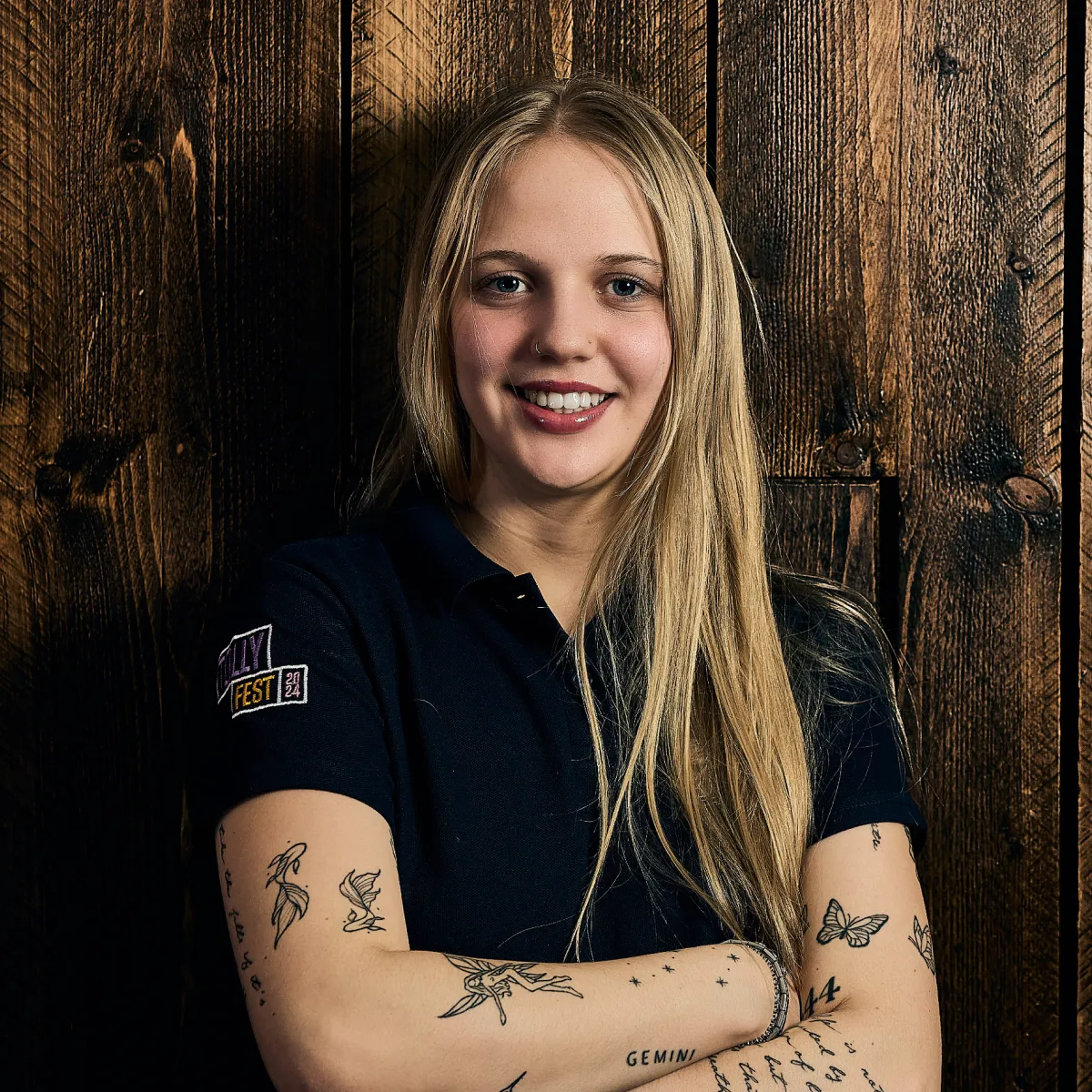 Smiling young woman with long blonde hair, crossed arms showing various black ink tattoos, wearing a black shirt standing in front of a dark wooden wall.