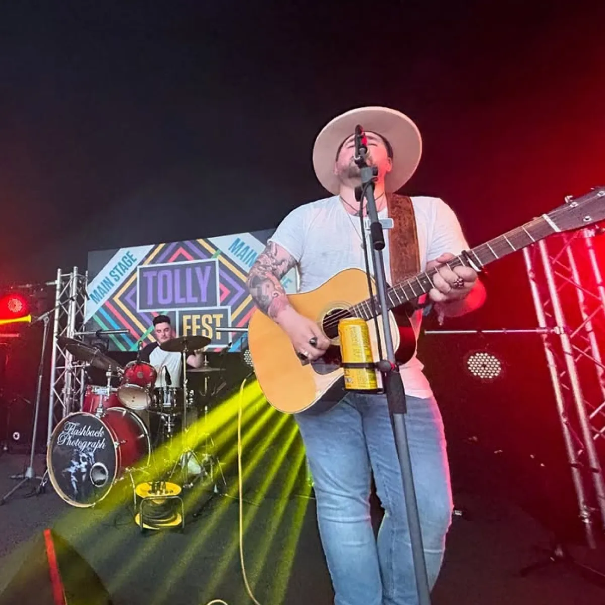 Musician with tattoos and hat playing acoustic guitar and singing on stage with drummer behind at Tolly Fest main stage.