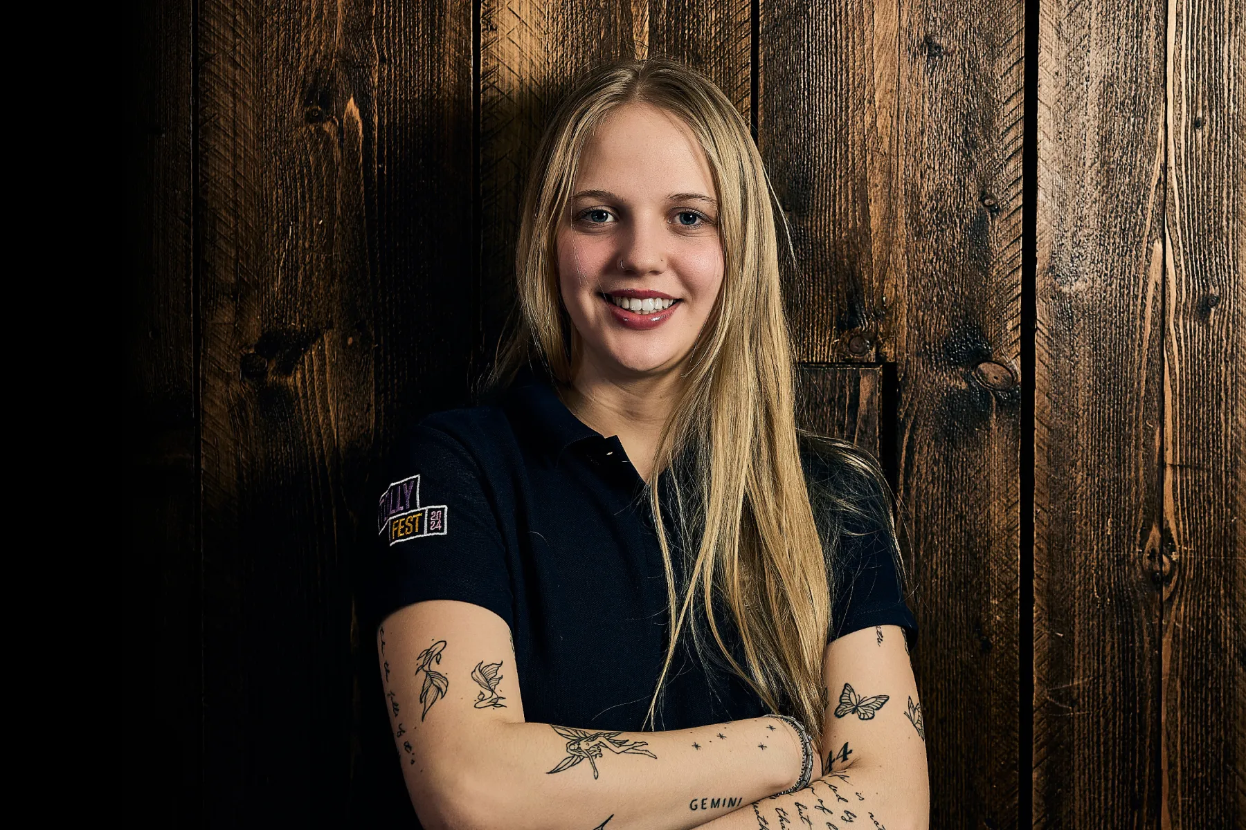 Young woman with long blonde hair and multiple tattoos on her arms smiling in front of a wooden wall.