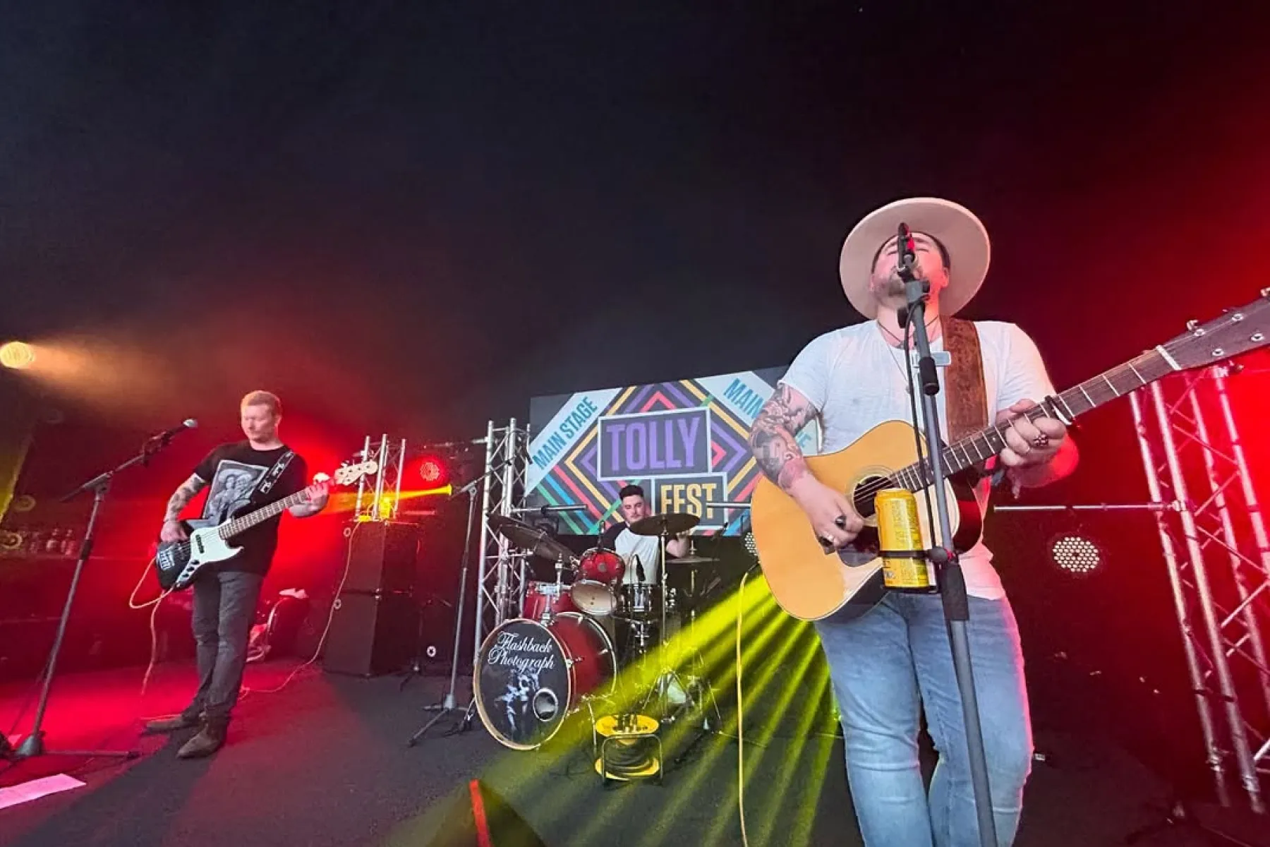 Three-member band performing on stage at Tolly Fest with red and yellow stage lights.