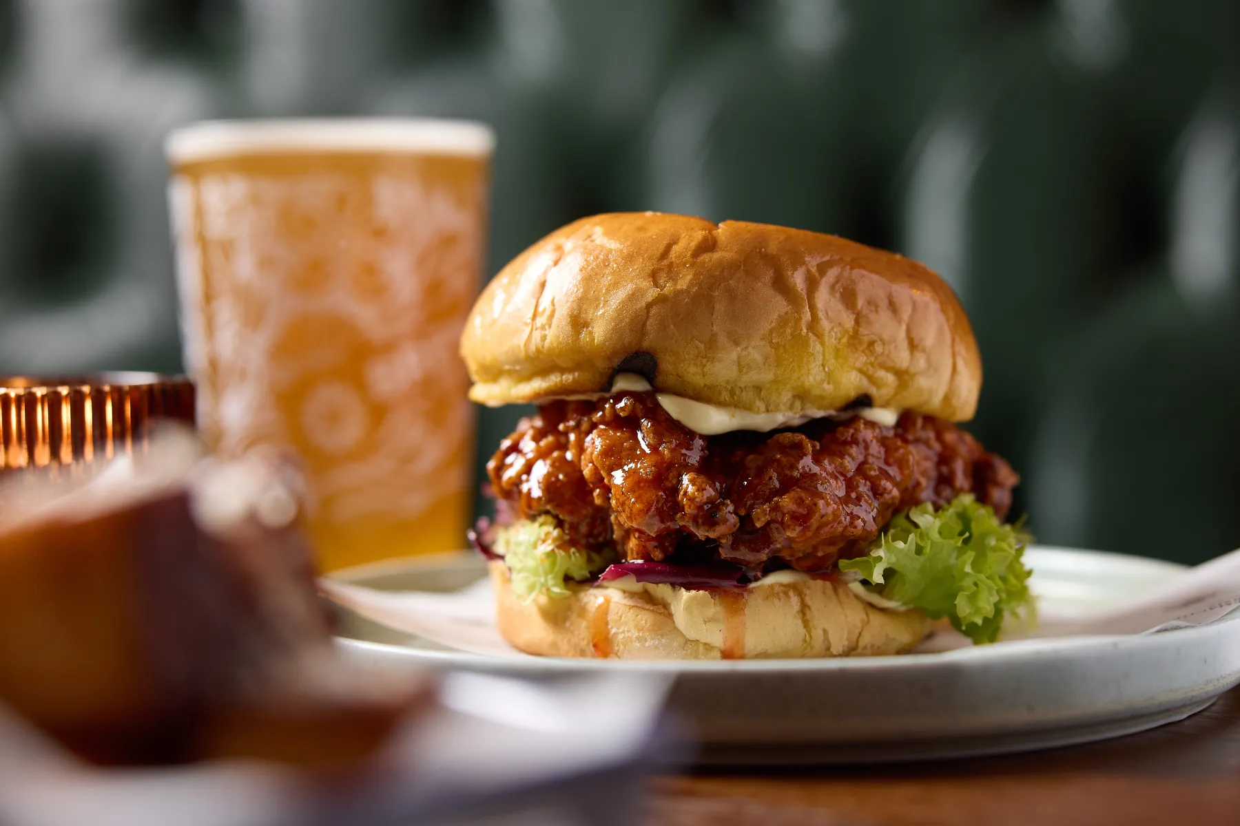 Spicy fried chicken sandwich with lettuce and mayonnaise on a soft bun, served on a white plate with a glass of beer in the background.